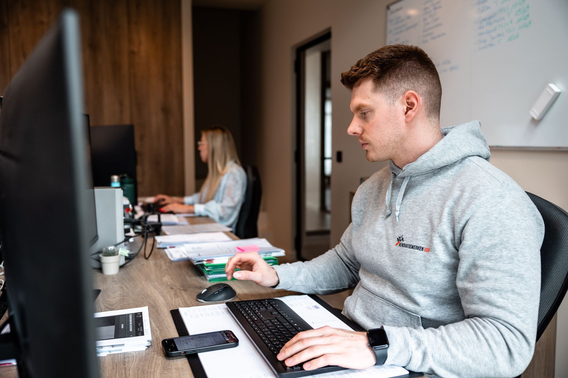Man in grijze hoodie werkt achter een computer, iemand anders op de achtergrond, kantooromgeving.
