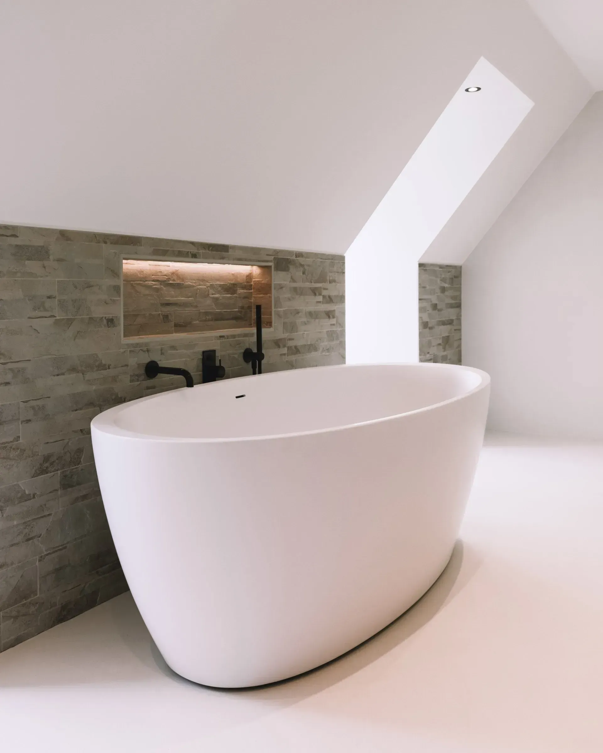 Wit ovaal bad in een moderne badkamer met stenen accentmuur en zwarte armaturen. Schuin plafond met natuurlijk licht.