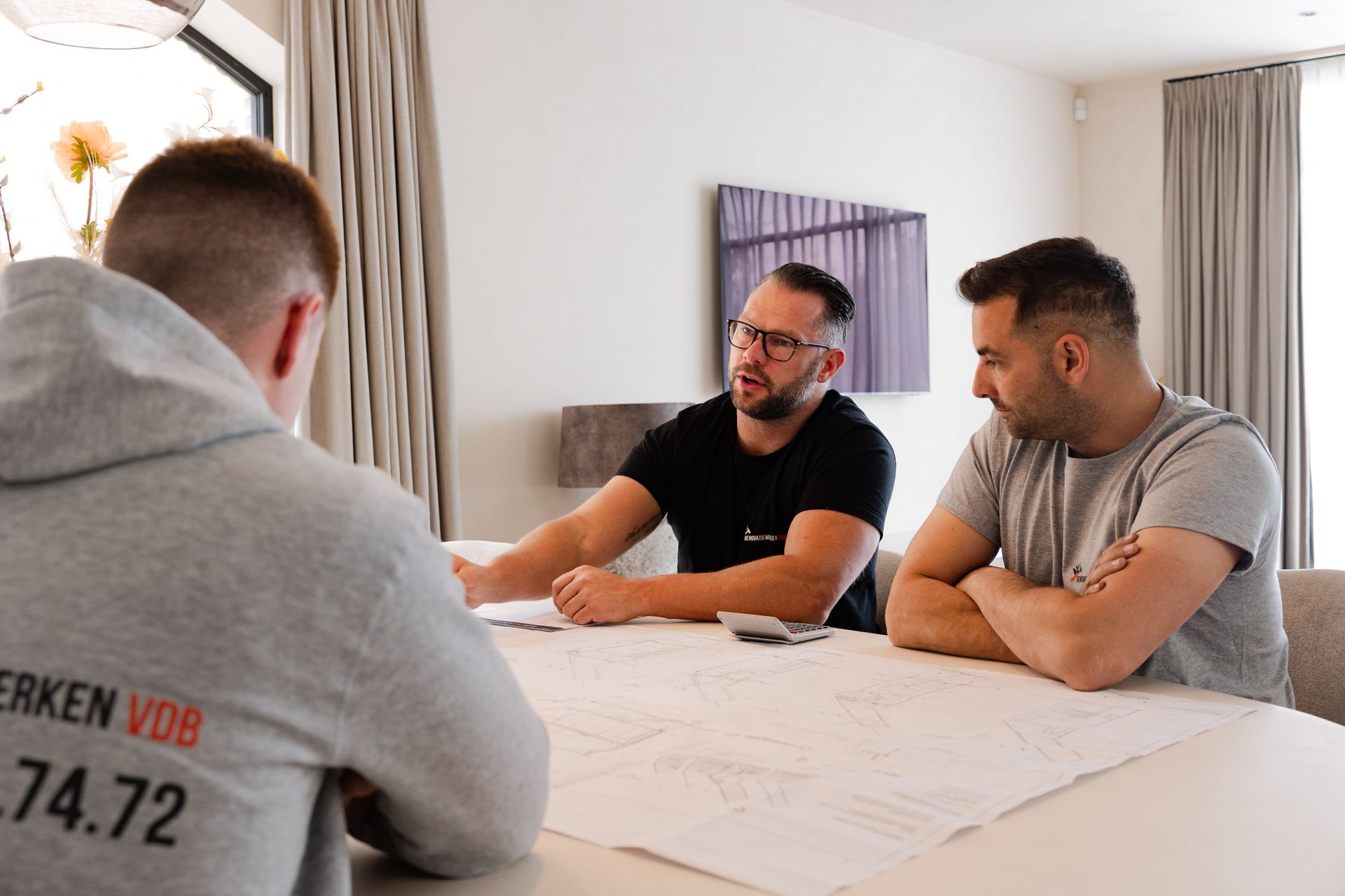 Drie mannen bespreken een plan aan een tafel. Eén wijst, anderen luisteren. Neutrale kamer, daglicht, geconcentreerd.