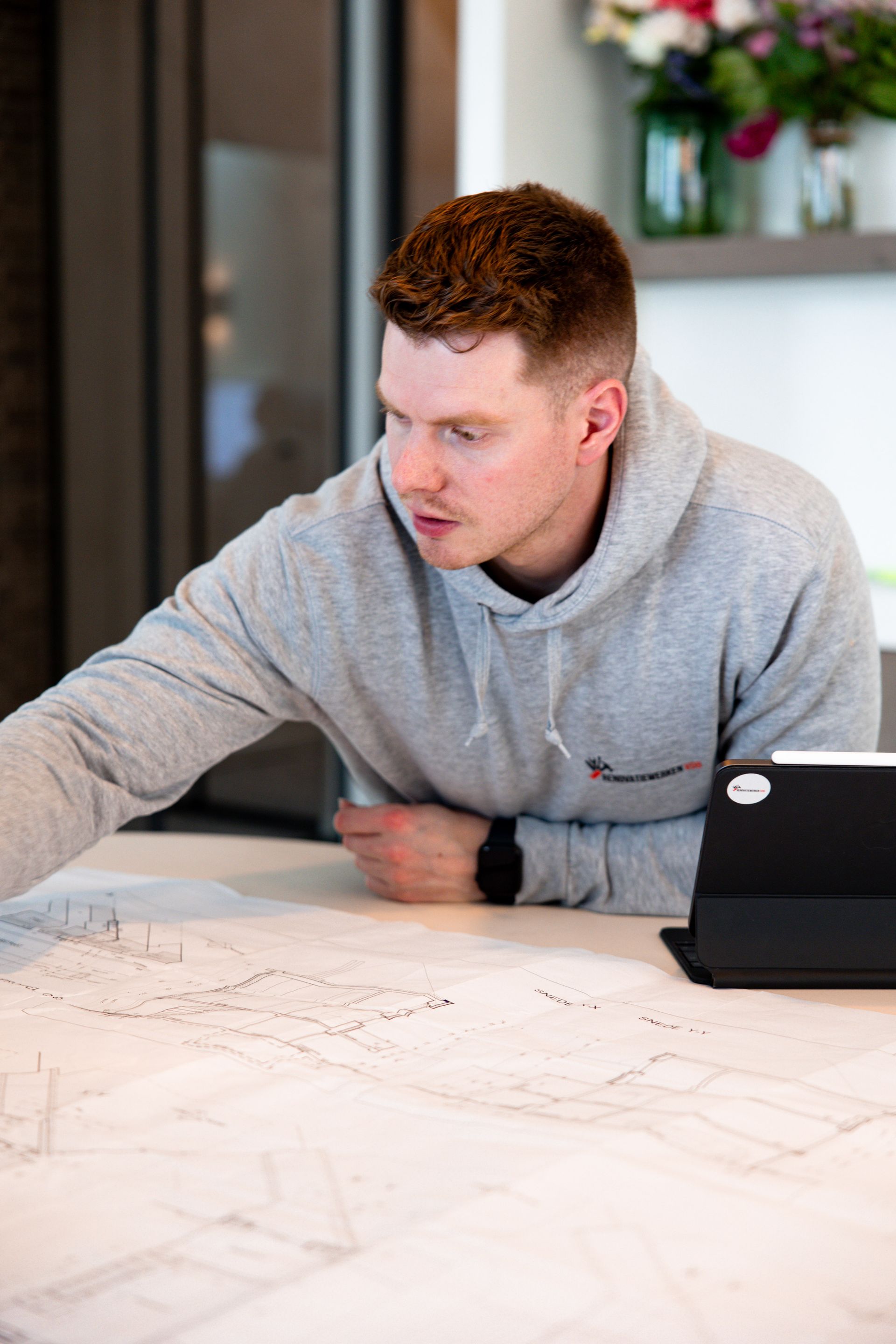 Man in grijze hoodie buigt zich over een grote blauwdruk en is gefocust. Tablet rechts.