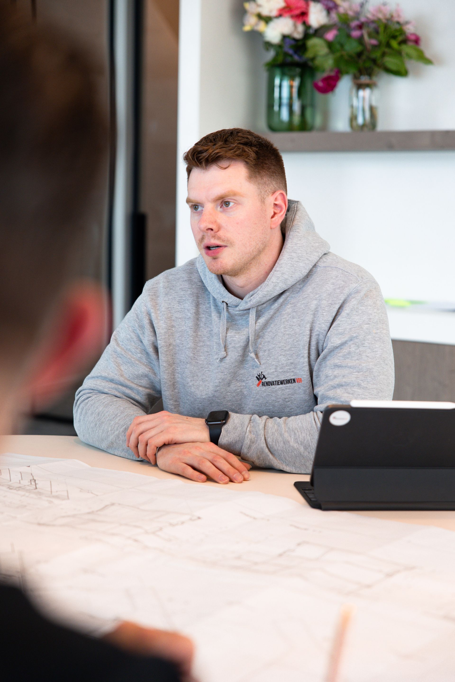 Man in grijze hoodie, bezorgd kijkend, zittend aan een tafel, pratend met iemand anders, bij een tablet en een blauwdruk.