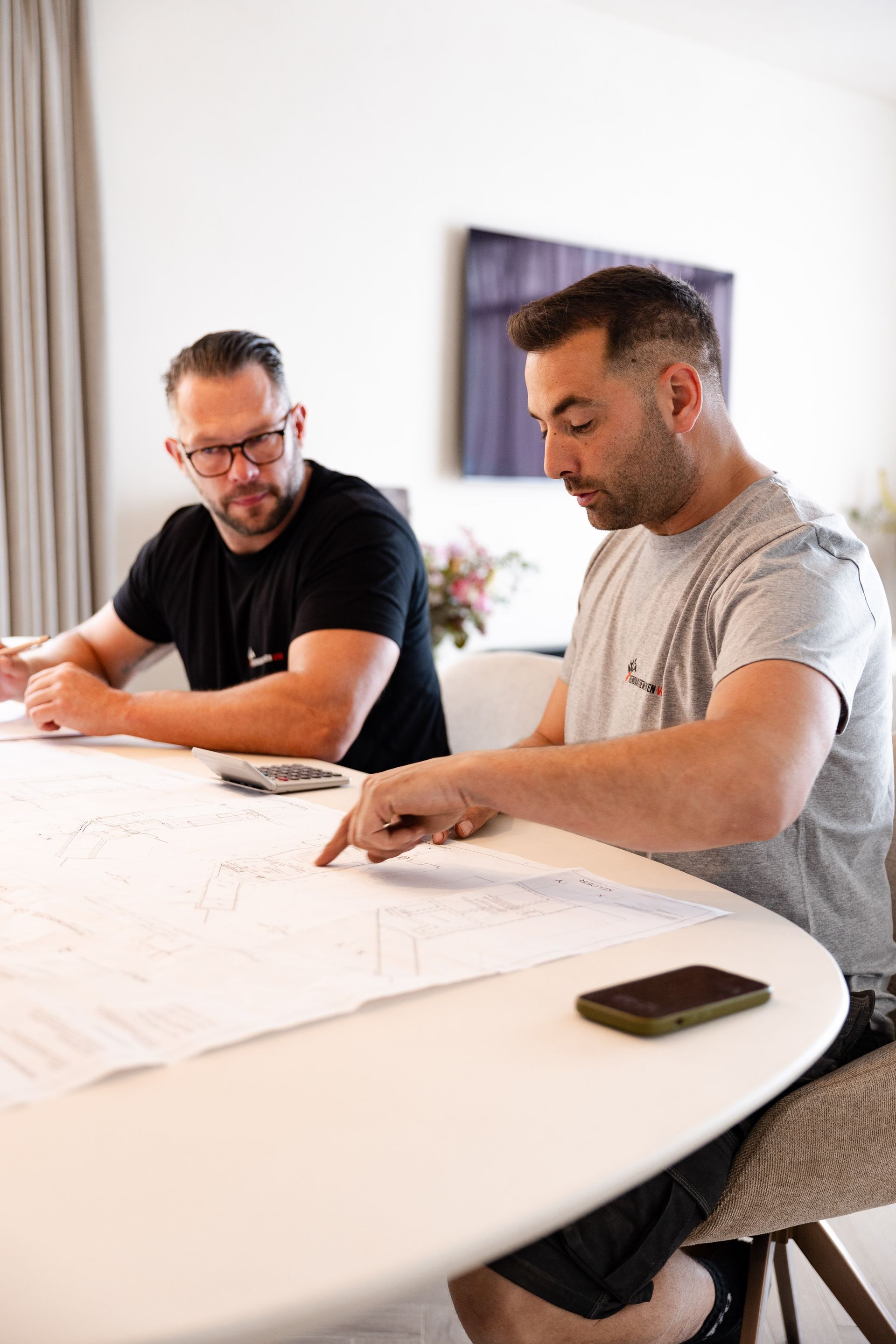 Twee mannen bekijken bouwtekeningen aan een witte tafel in een felverlichte kamer; een van hen wijst naar de plannen.