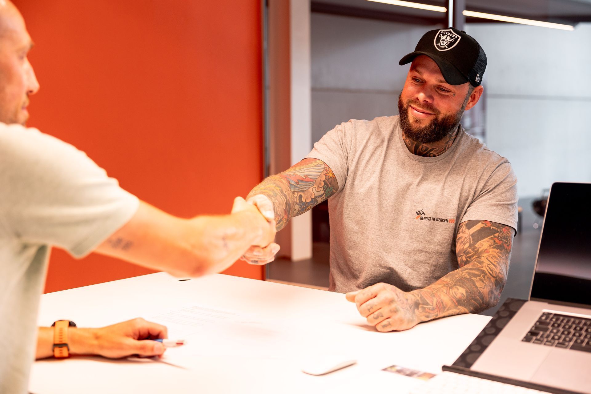 Twee mannen schudden elkaar de hand aan een bureau, een van hen met tatoeages en een pet op, in een modern kantoor.