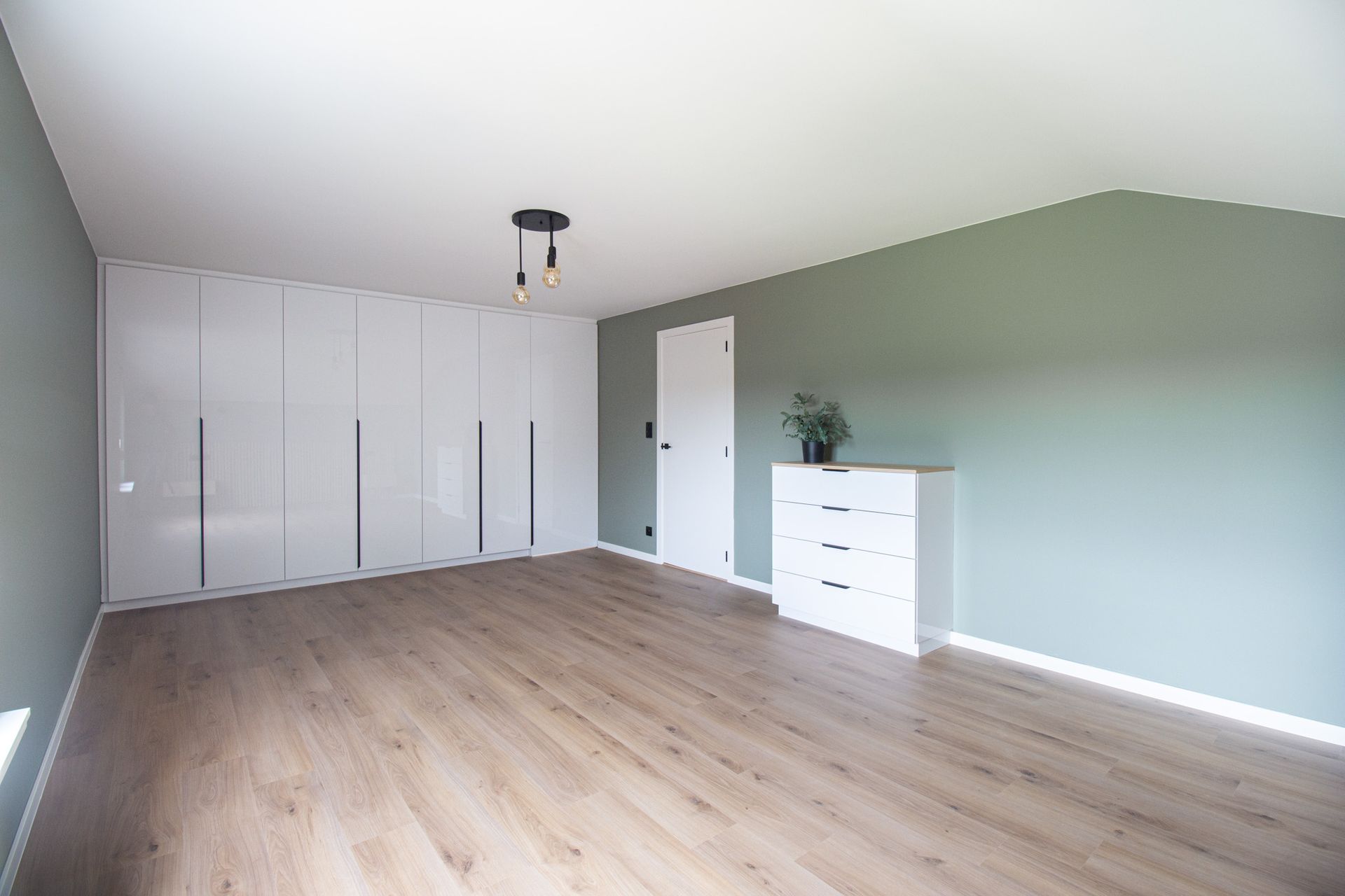 Lege slaapkamer met witte inbouwkast, lichtgroene muur, witte ladekast en houten vloer.