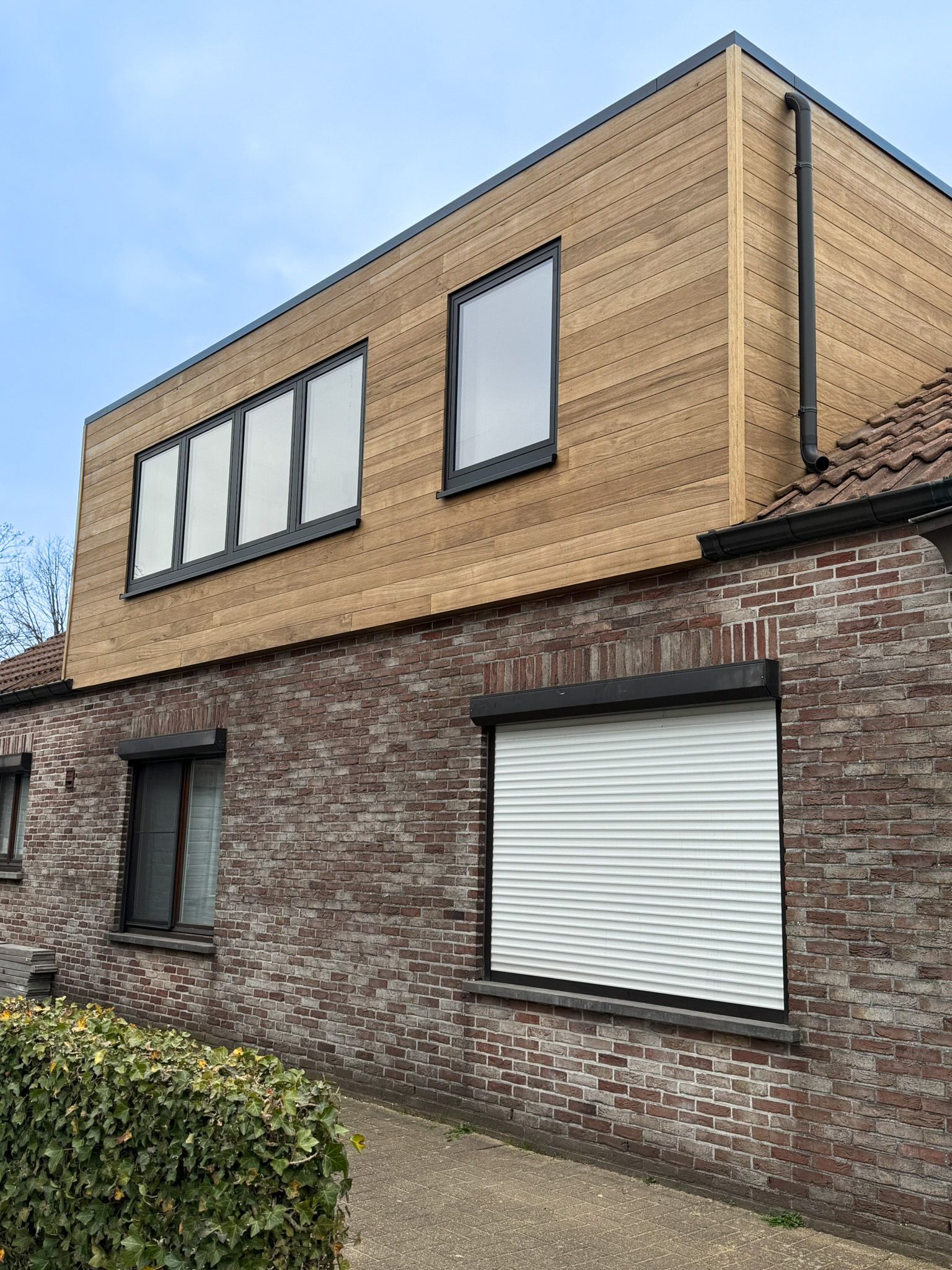 Gebouw met twee verdiepingen: bakstenen onderbouw, houten bovenbouw. ​​Ramen met zwarte kozijnen, rolluiken.