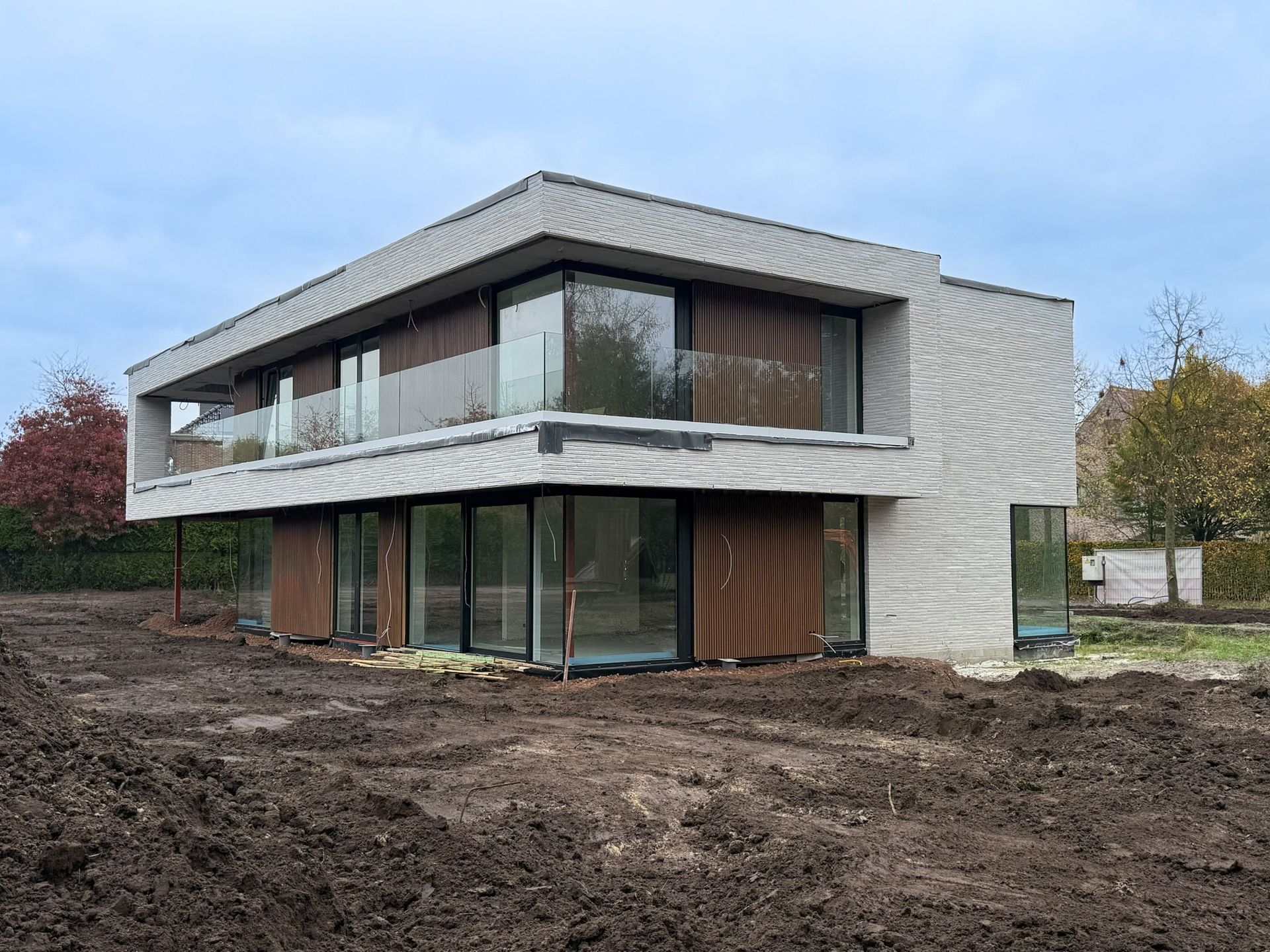 Modern huis met twee verdiepingen in aanbouw; betonnen gevel, glazen ramen, houten accenten en modderige grond.