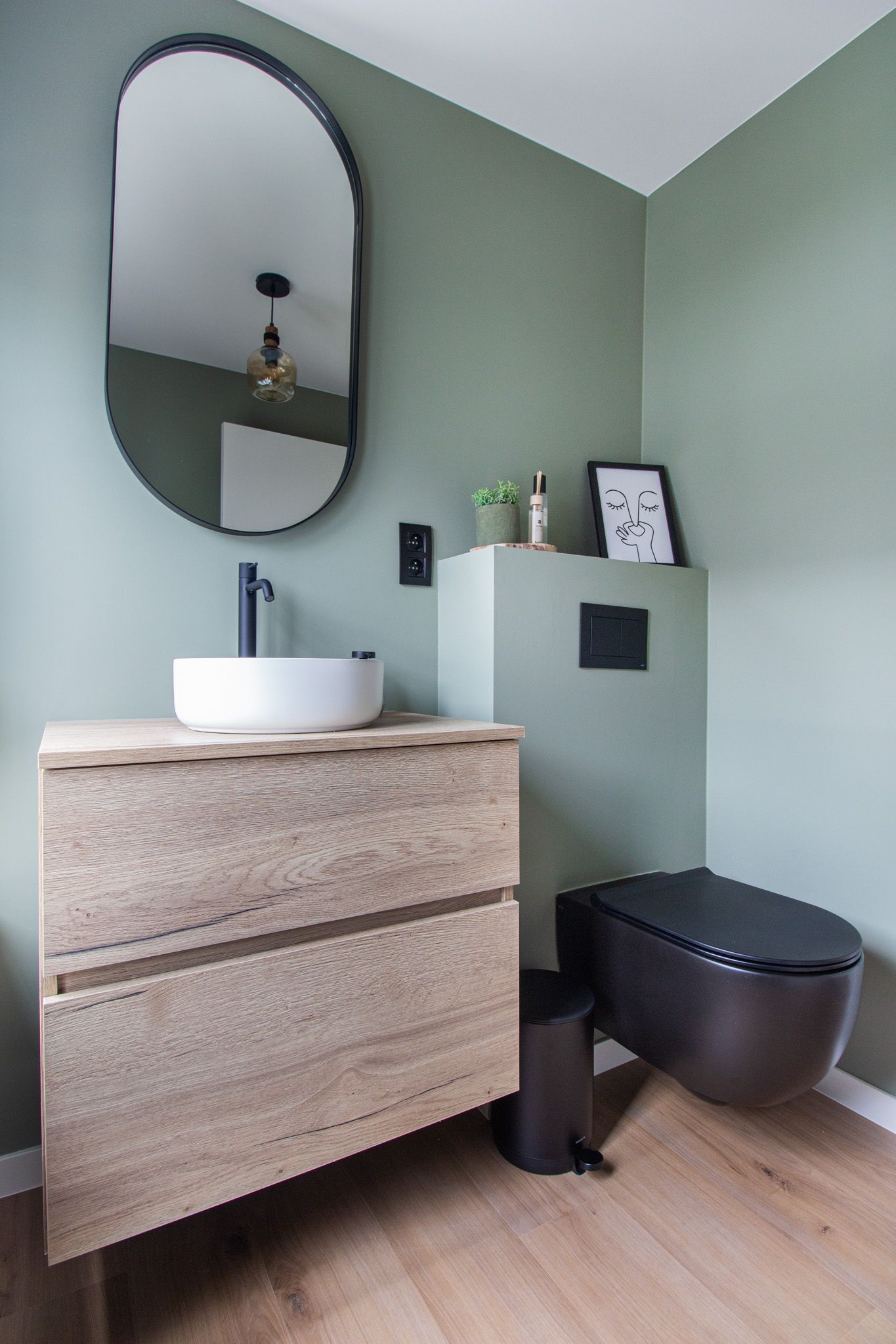 Badkamer met saliegroene muren, houten wastafelmeubel met witte wastafel, zwart toilet en ovale spiegel.