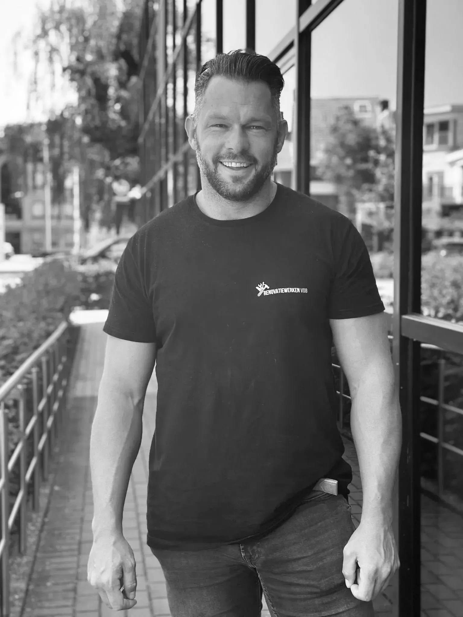 Lachende man, gekleed in een donker T-shirt en spijkerbroek, staat buiten een gebouw.