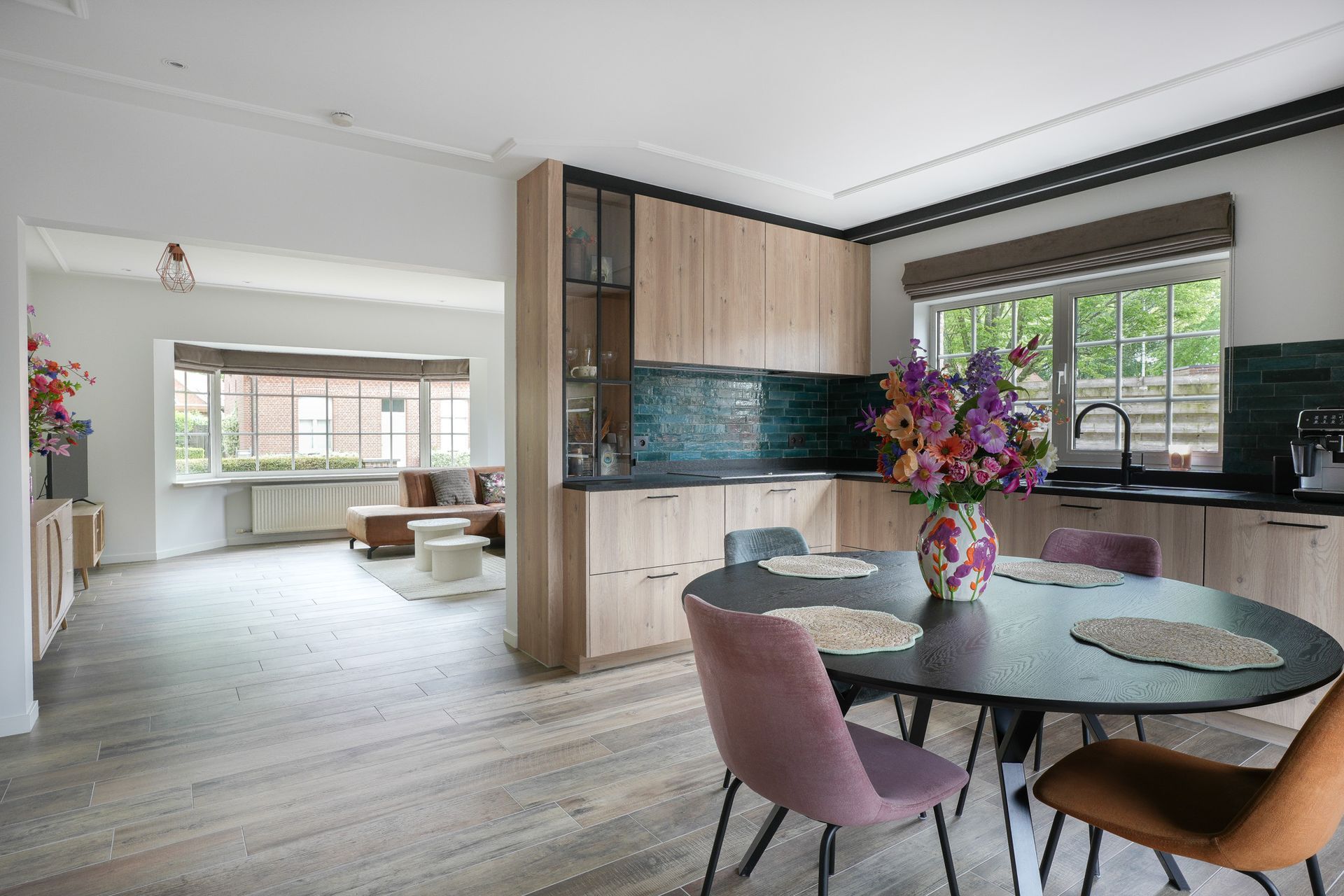 Open keuken met een ronde tafel en kleurrijke stoelen, lichte houten kasten en een woongedeelte op de achtergrond.