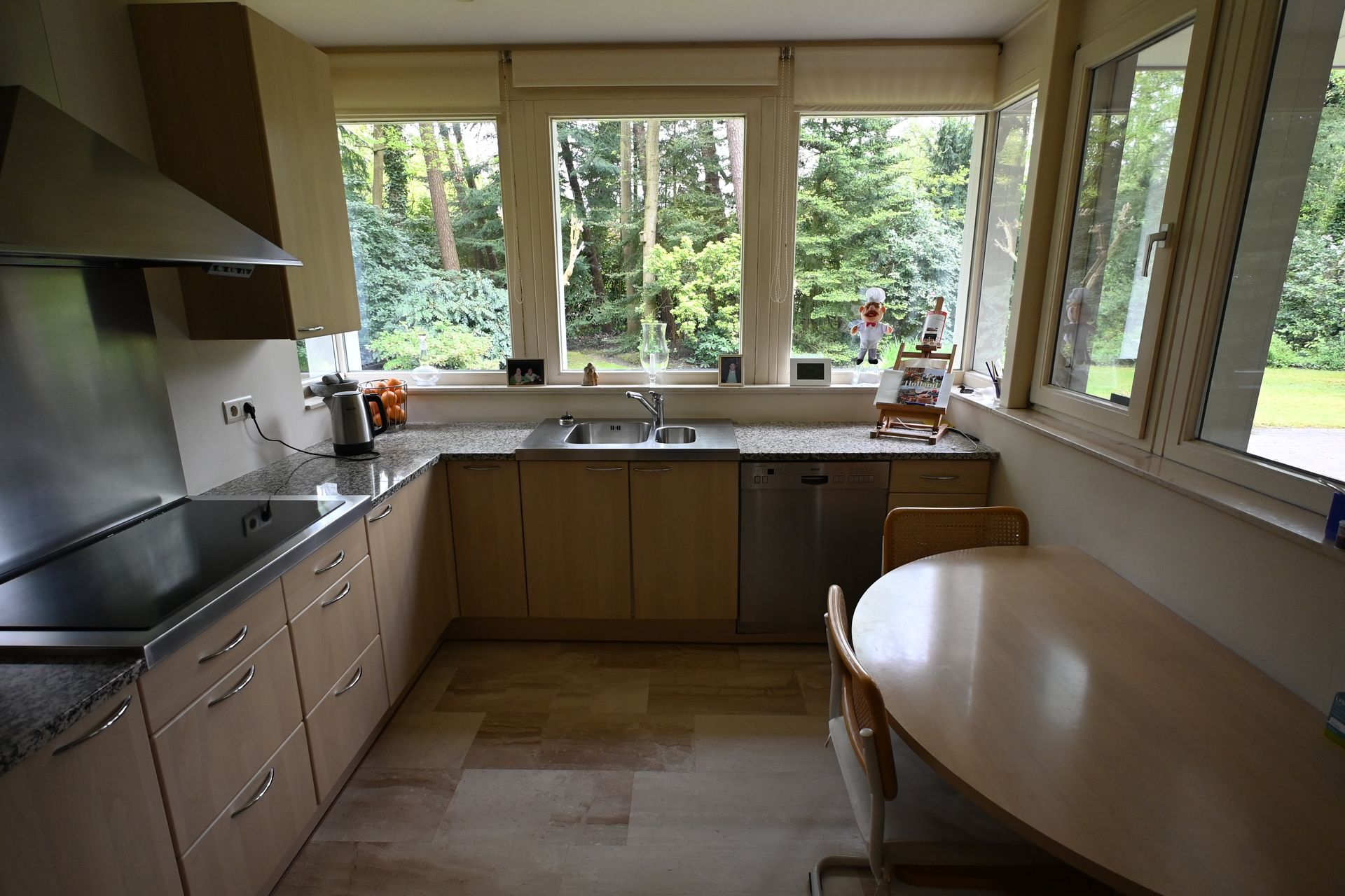 Keuken met lichte houten kasten, roestvrijstalen apparatuur, een spoelbak en een eettafel naast grote ramen met uitzicht op een groen landschap.