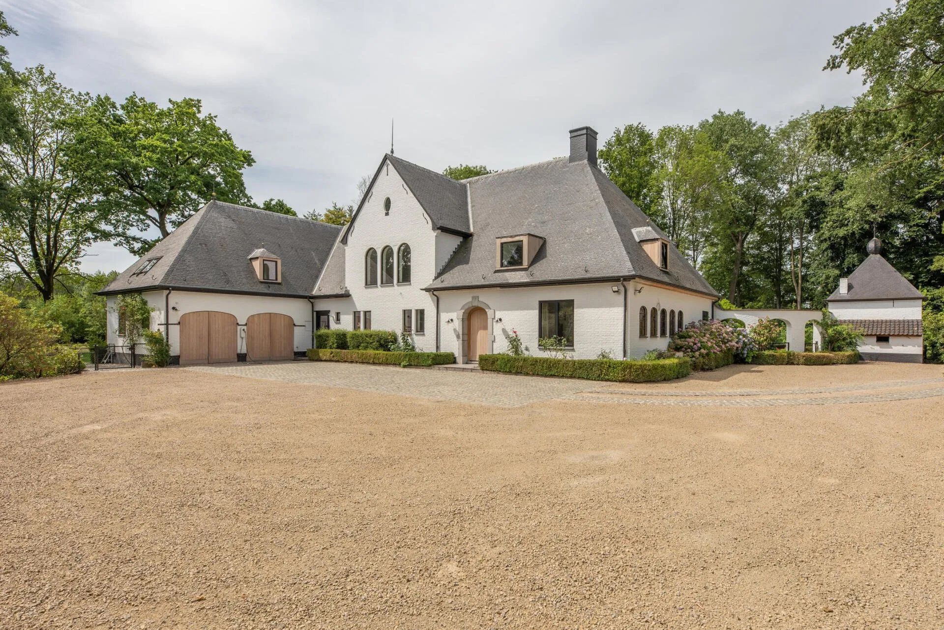 Wit huis met een grindpad, geflankeerd door bomen en een klein bijgebouw.