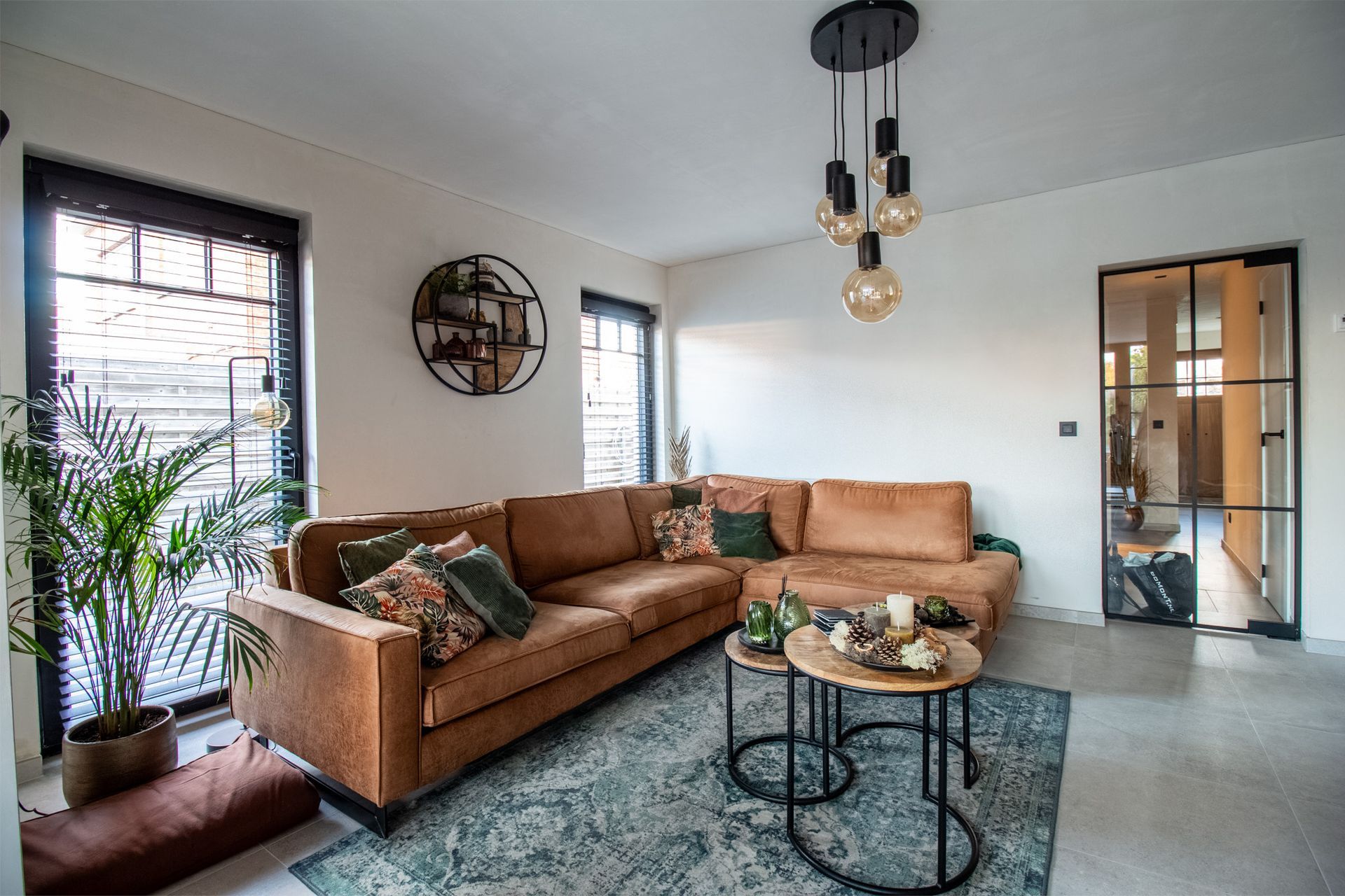 Woonkamer met bruine hoekbank, vloerkleed en twee kleine salontafels. Zwarte accenten en hanglampen.