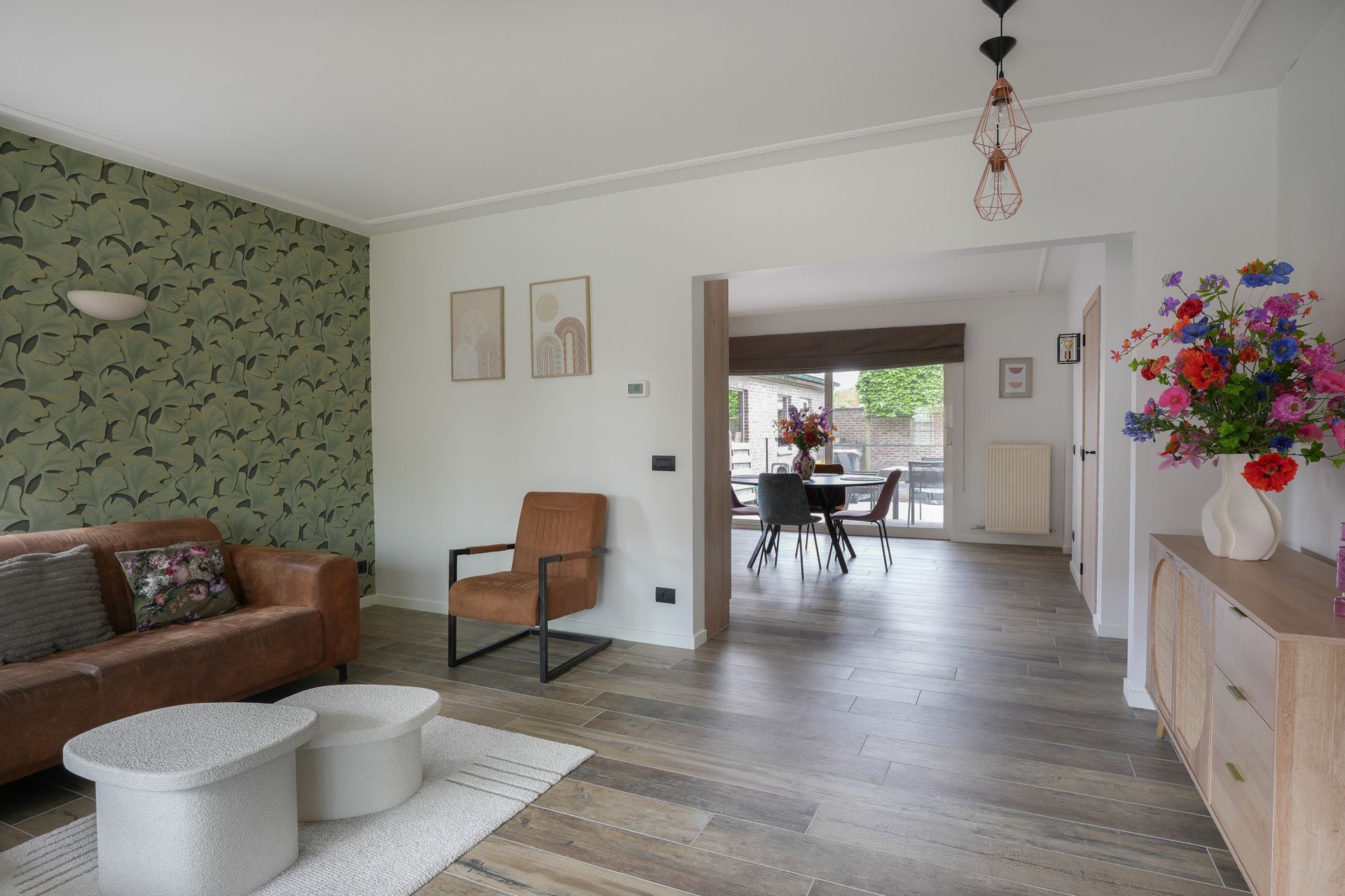 Woonkamer met bruine bank, behang met patroon, zichtbare eethoek en bloemstuk.