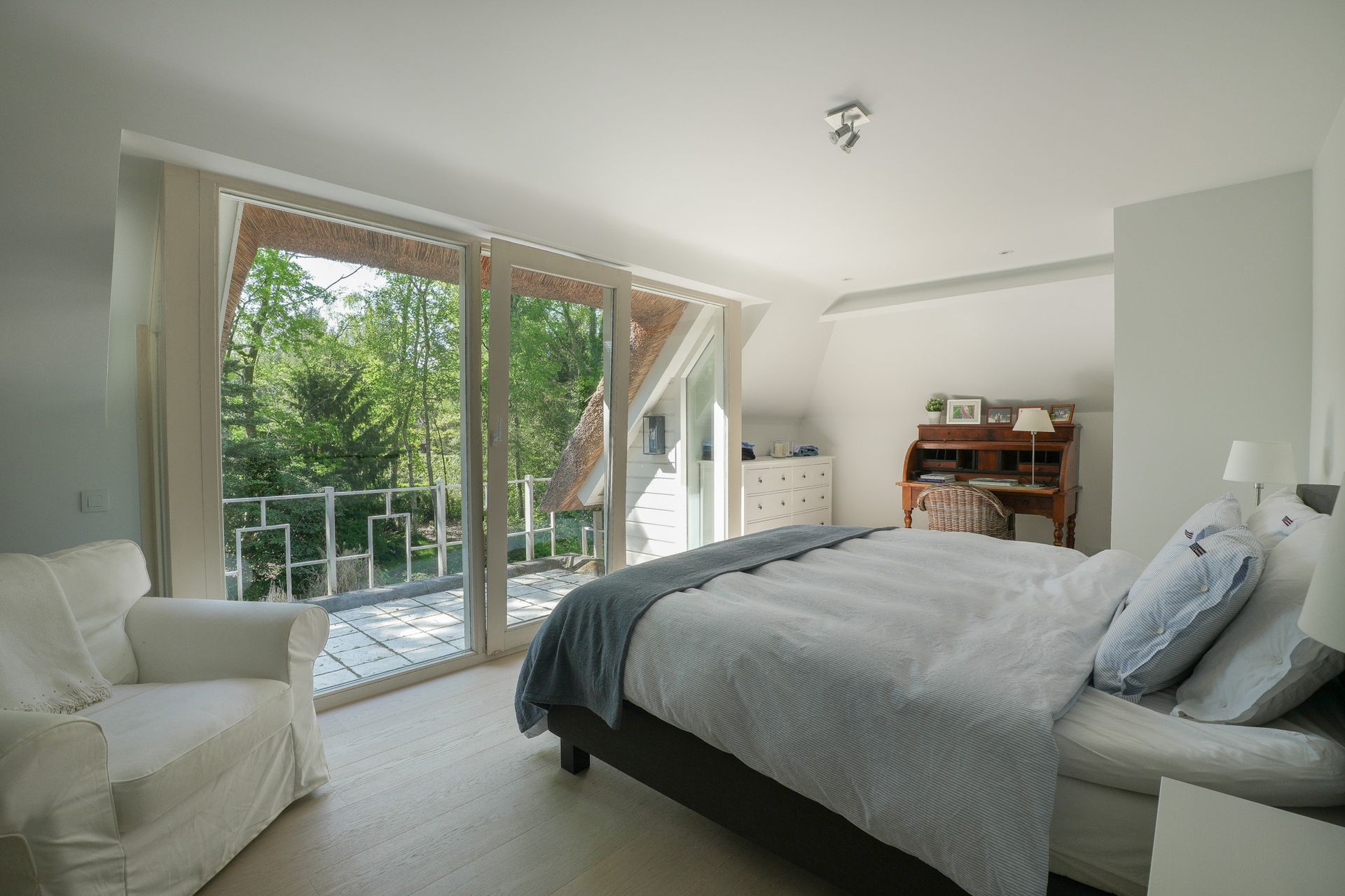 Slaapkamer met grote ramen met uitzicht op de bomen, wit bed, fauteuil.