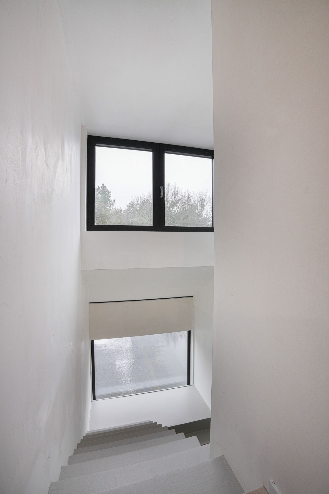 Trappenhuis met witte muren, een raam met zwarte kozijnen boven en nog een lager gelegen raam, zichtbare treden en bewolkt uitzicht.