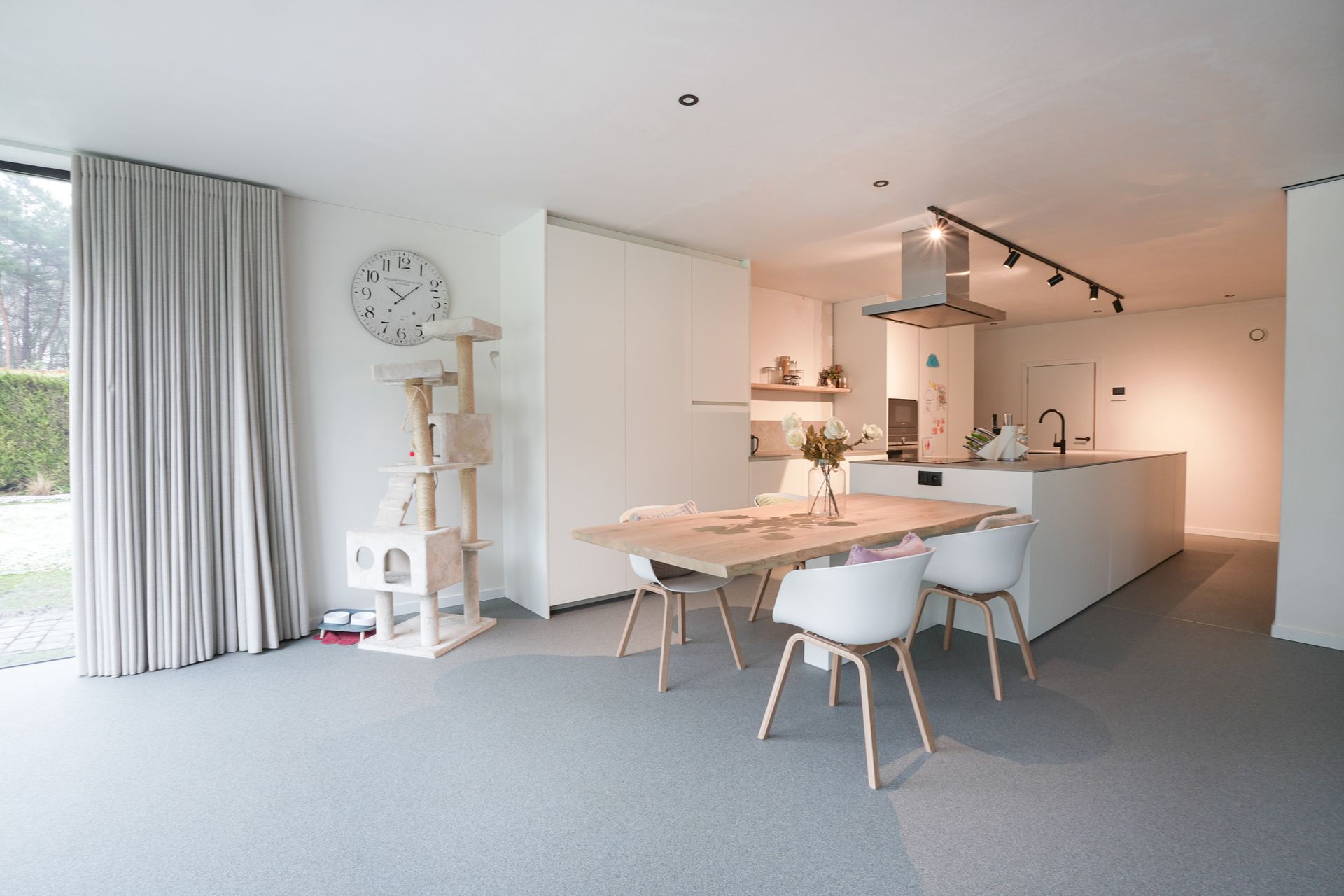 Moderne open keuken en eetkamer. Witte muren, lichtgekleurd meubilair, een krabpaal en een groot raam.