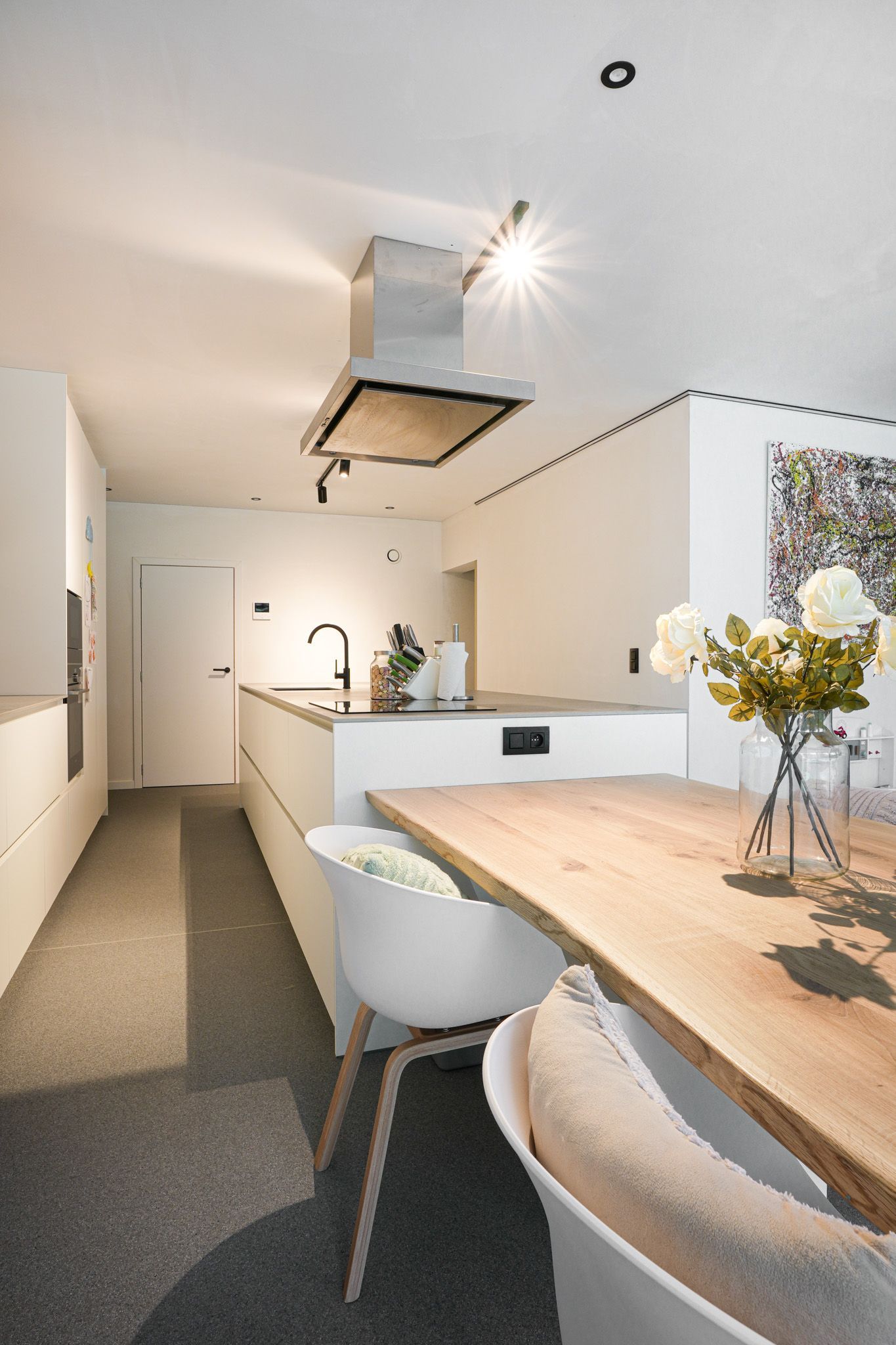 Moderne witte keuken met kookeiland, houten tafel en witte stoelen, verlicht door spots.