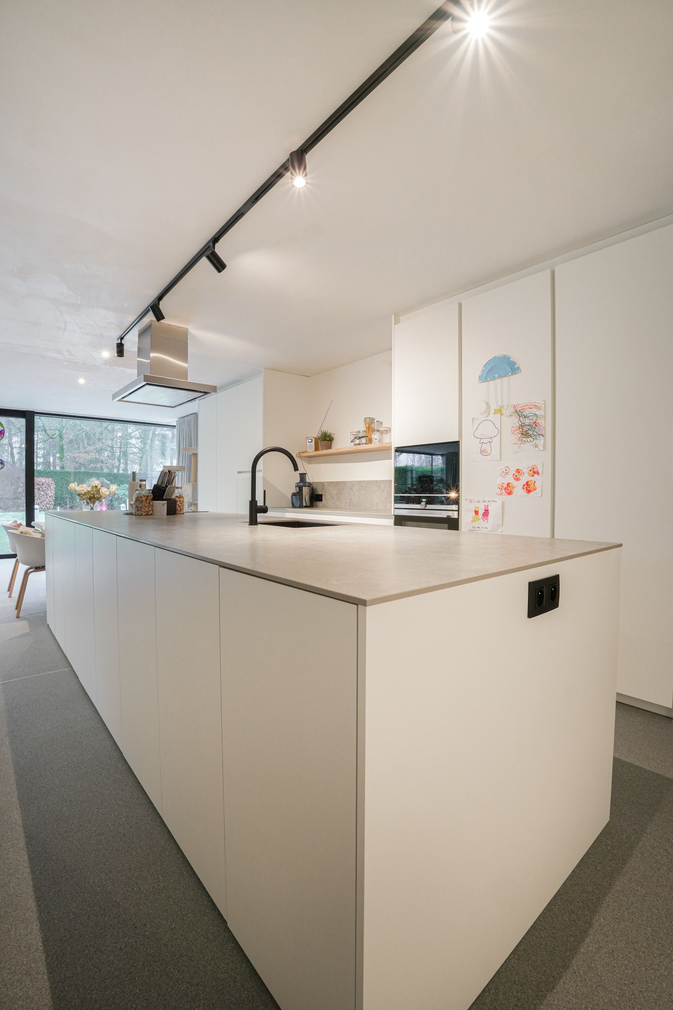 Moderne witte keuken met kookeiland, railsverlichting, zwarte kraan, inbouwoven en raam met uitzicht op het groen.
