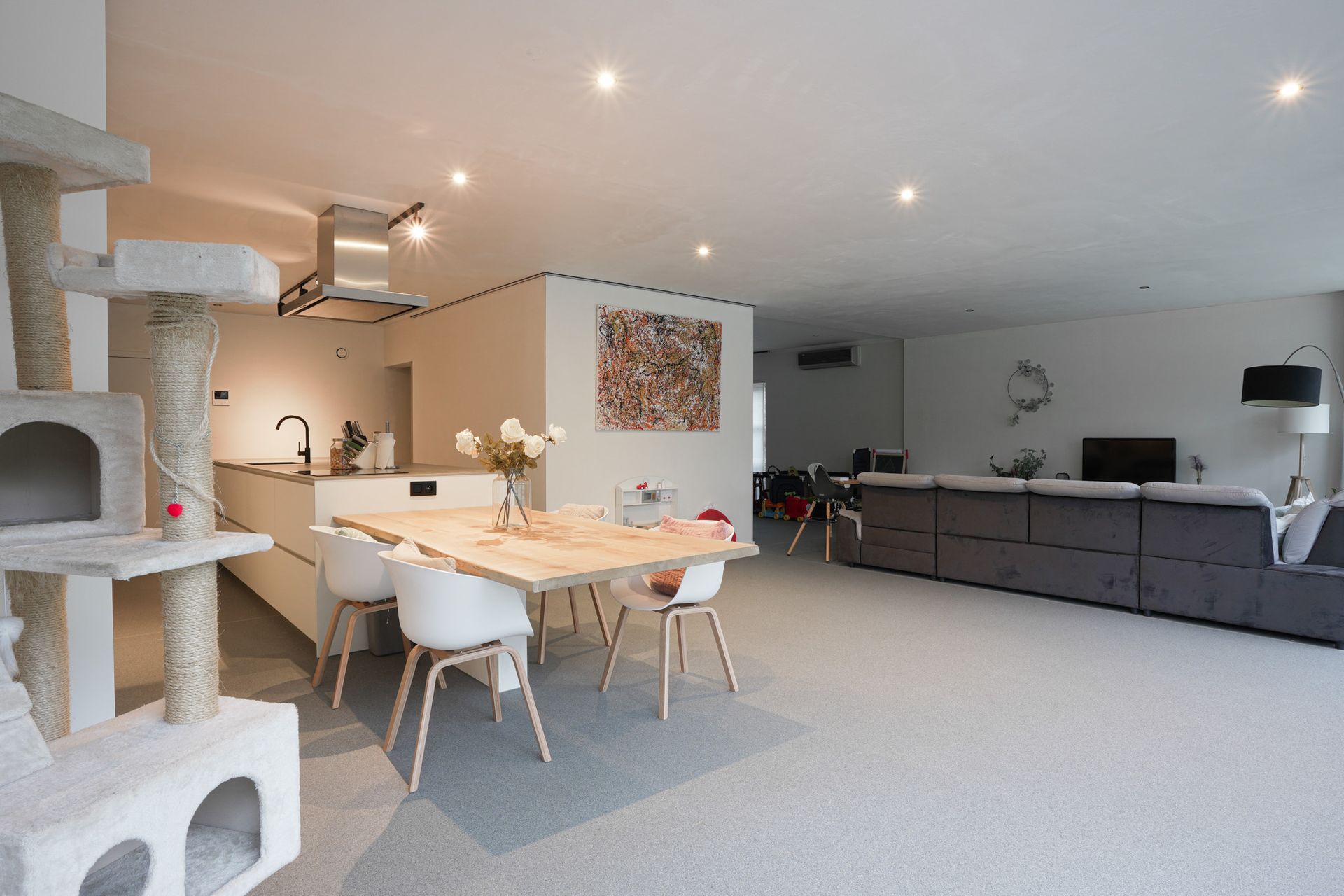 Open leefruimte: keuken, eetkamer en woonkamer. Lichtgekleurd meubilair, grijs tapijt, grote kattenboom.