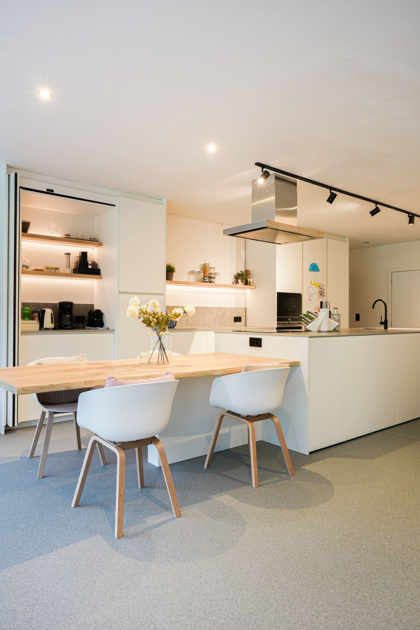 Moderne witte keuken met een tafel met houten blad, witte stoelen, open planken en ingebouwde apparatuur.
