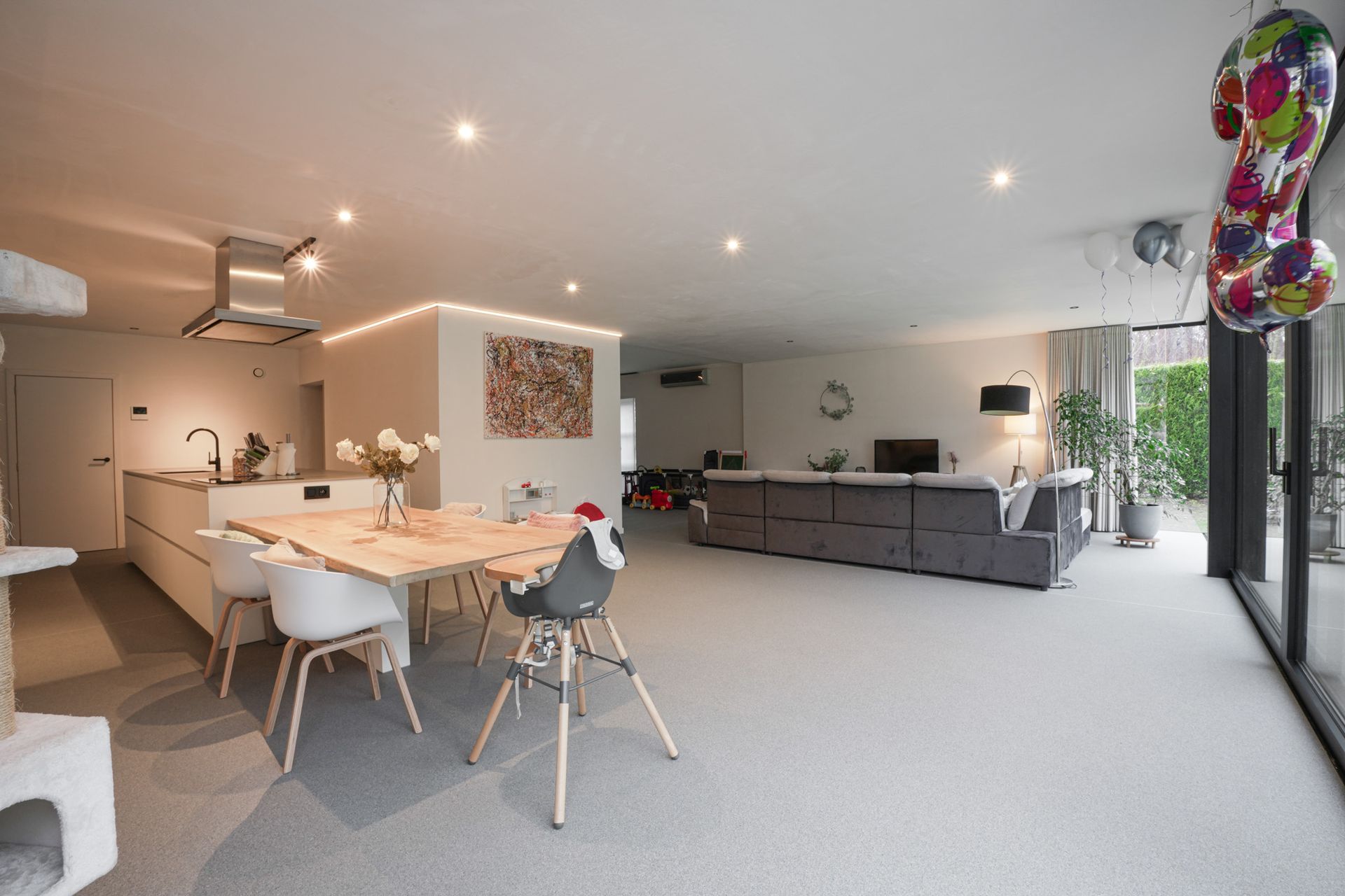 Open keuken en woonkamer met eettafel, grijze bank en grote ramen.