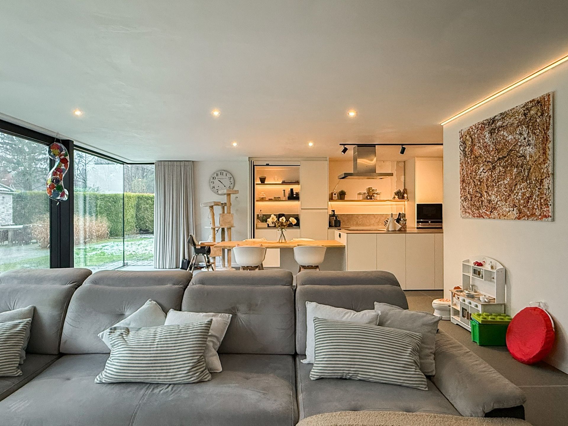 Moderne, open woonkamer met een grijze bank, keuken en groot raam met uitzicht naar buiten.