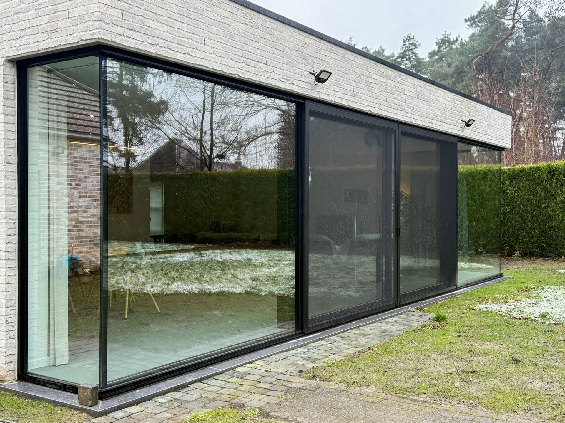 Modern huis met grote glazen ramen en schuifdeuren, gazon en bakstenen muur.