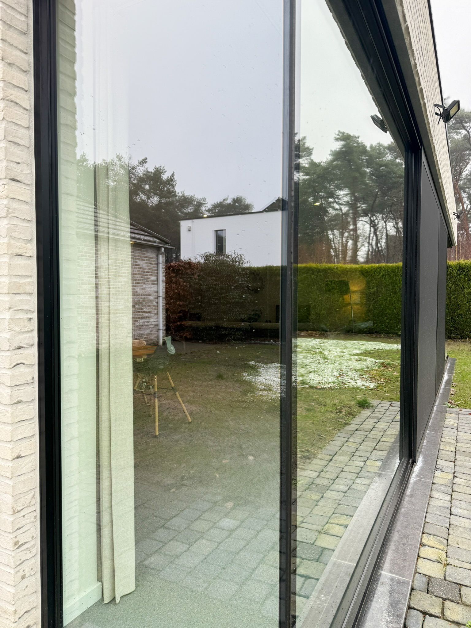 Groot raam met uitzicht op een achtertuin met bomen, gras, een wit huis en een stenen terras.