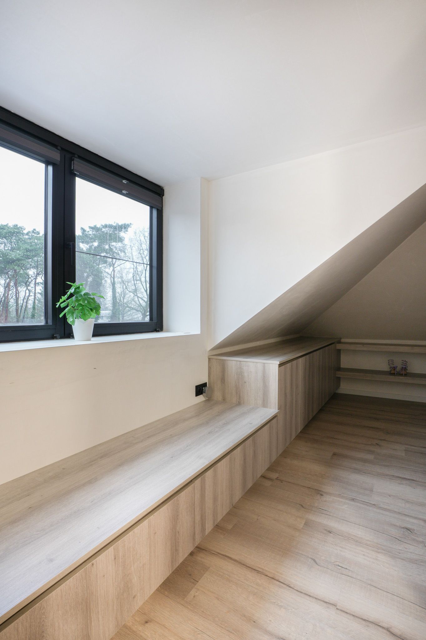 Zolderinterieur met houten bank, ingebouwde opbergruimte, raam en schuin plafond.
