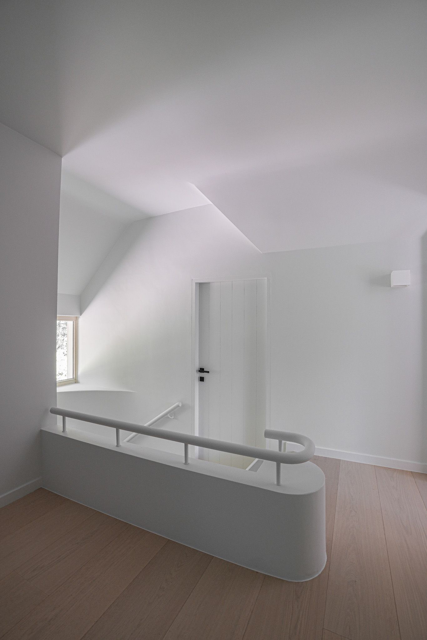 Interieur van een kamer met witte muren, een trapleuning, deur en raam, en een lichtgekleurde houten vloer.