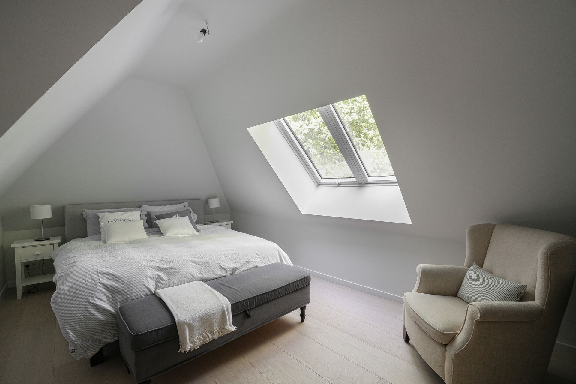 Slaapkamer met schuin plafond, bed, fauteuil en dakraam. Witte muren en meubels, zacht licht.