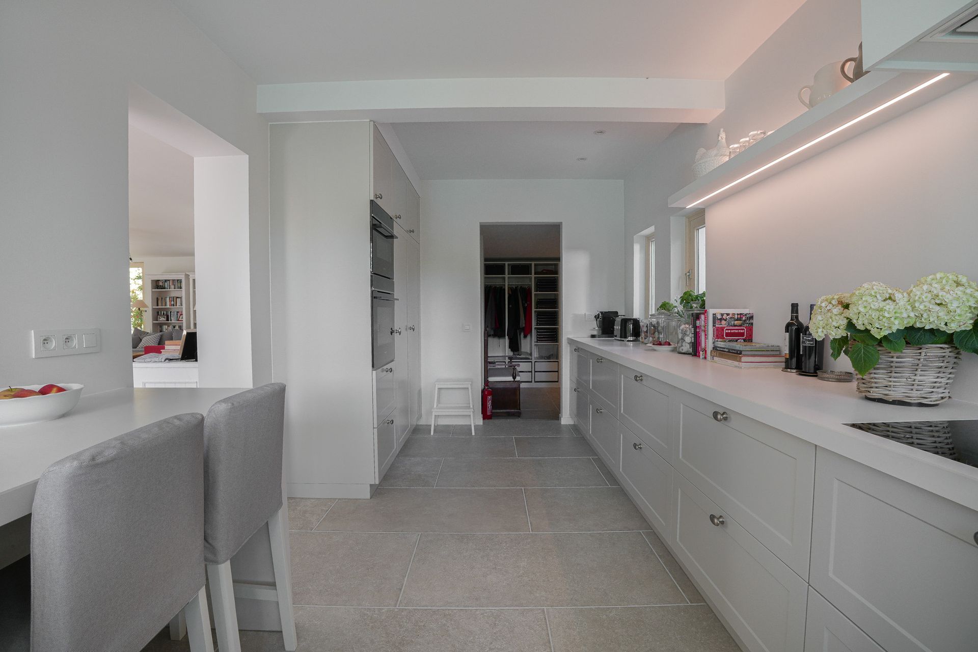 Een lichte, moderne keuken met witte kasten, stenen vloeren en een lang aanrechtblad.