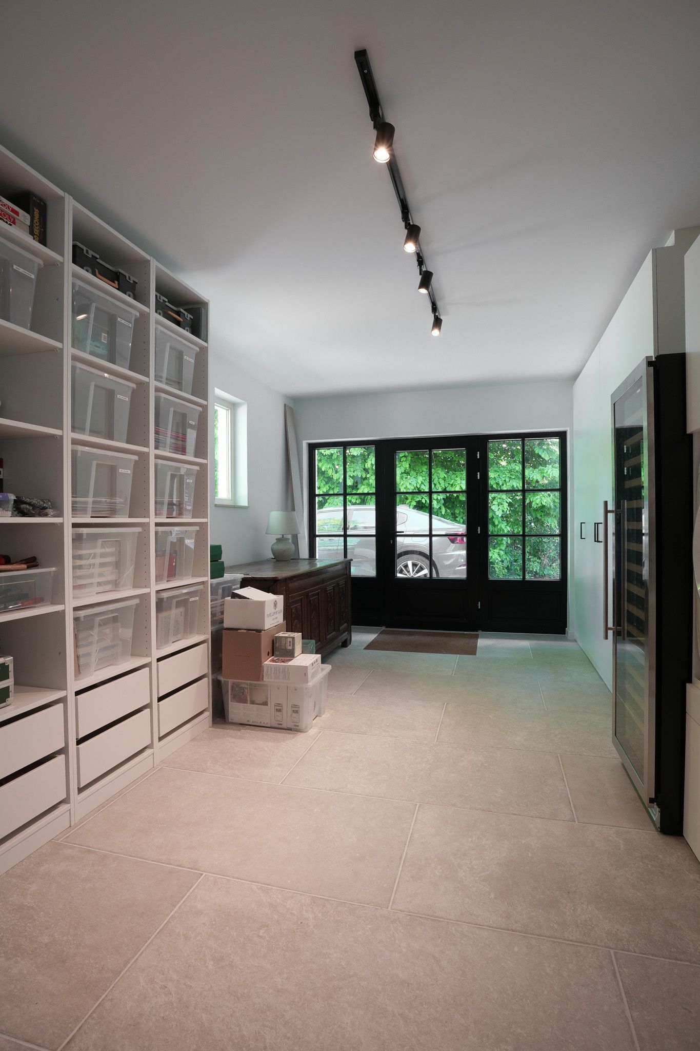 Lange, lichte kamer met ingebouwde witte planken, doorzichtige prullenbakken en een zwarte glazen deur.