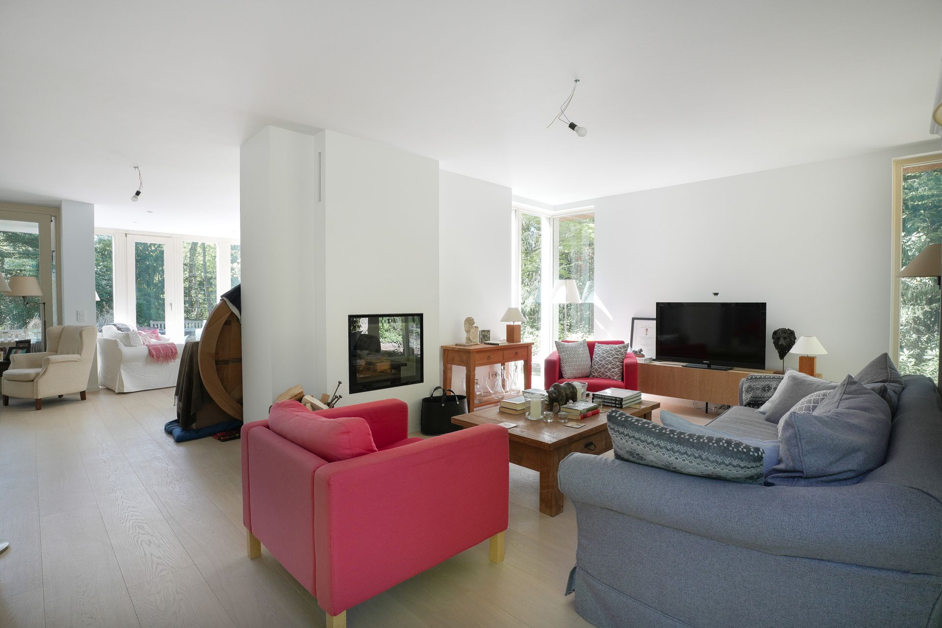Ruime woonkamer met open haard, twee rode stoelen, een blauwe bank en een grote televisie.
