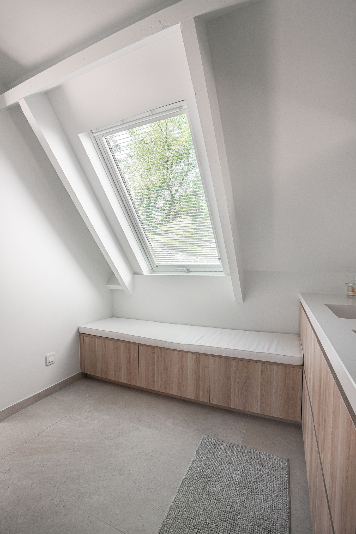 Een lichte badkamerhoek met een schuin raam, een houten bankje en een aanrechtblad van natuursteen.