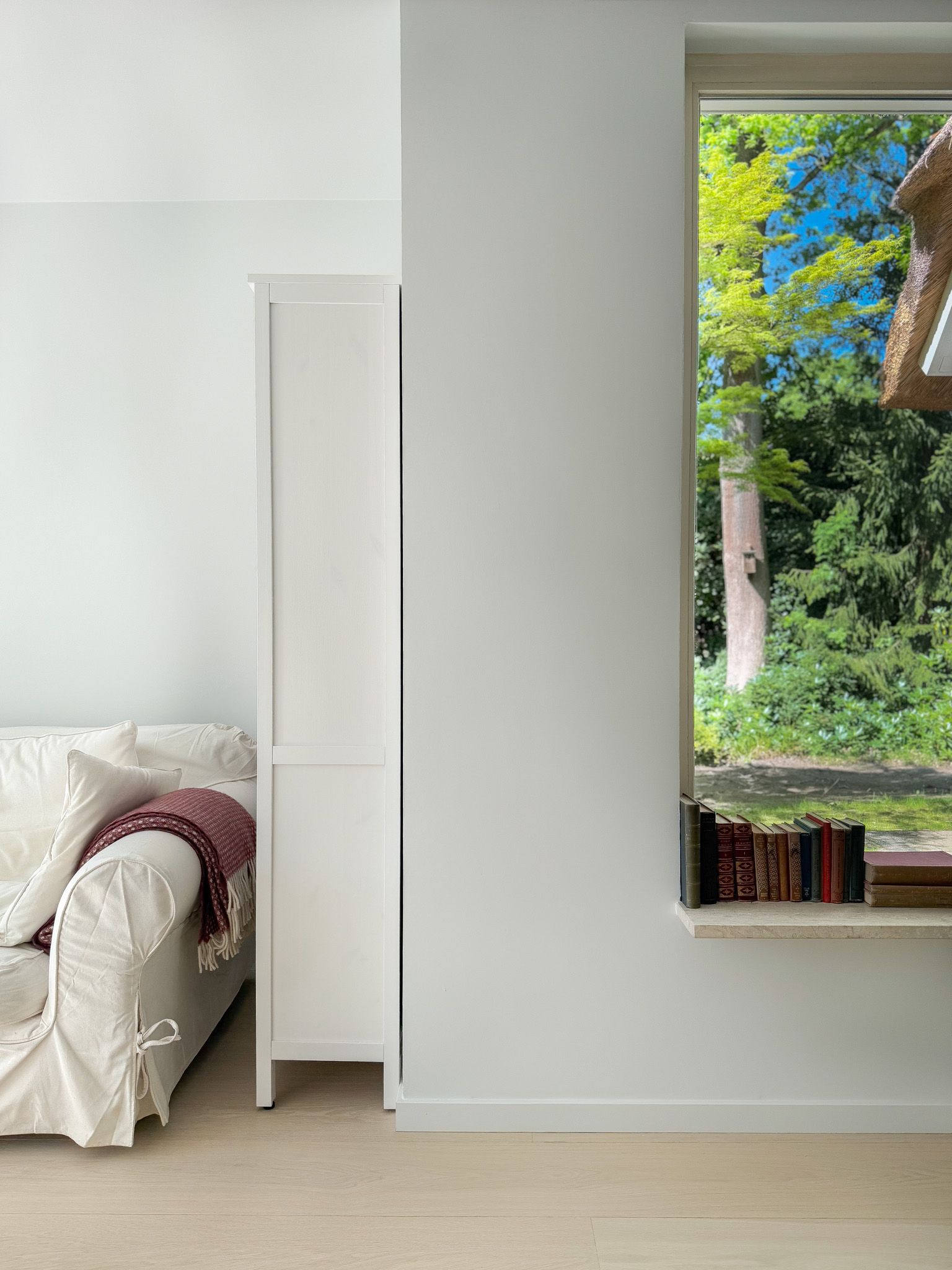 Witte muur, bank met deken, boekenkast, raam met uitzicht op bomen en boeken.