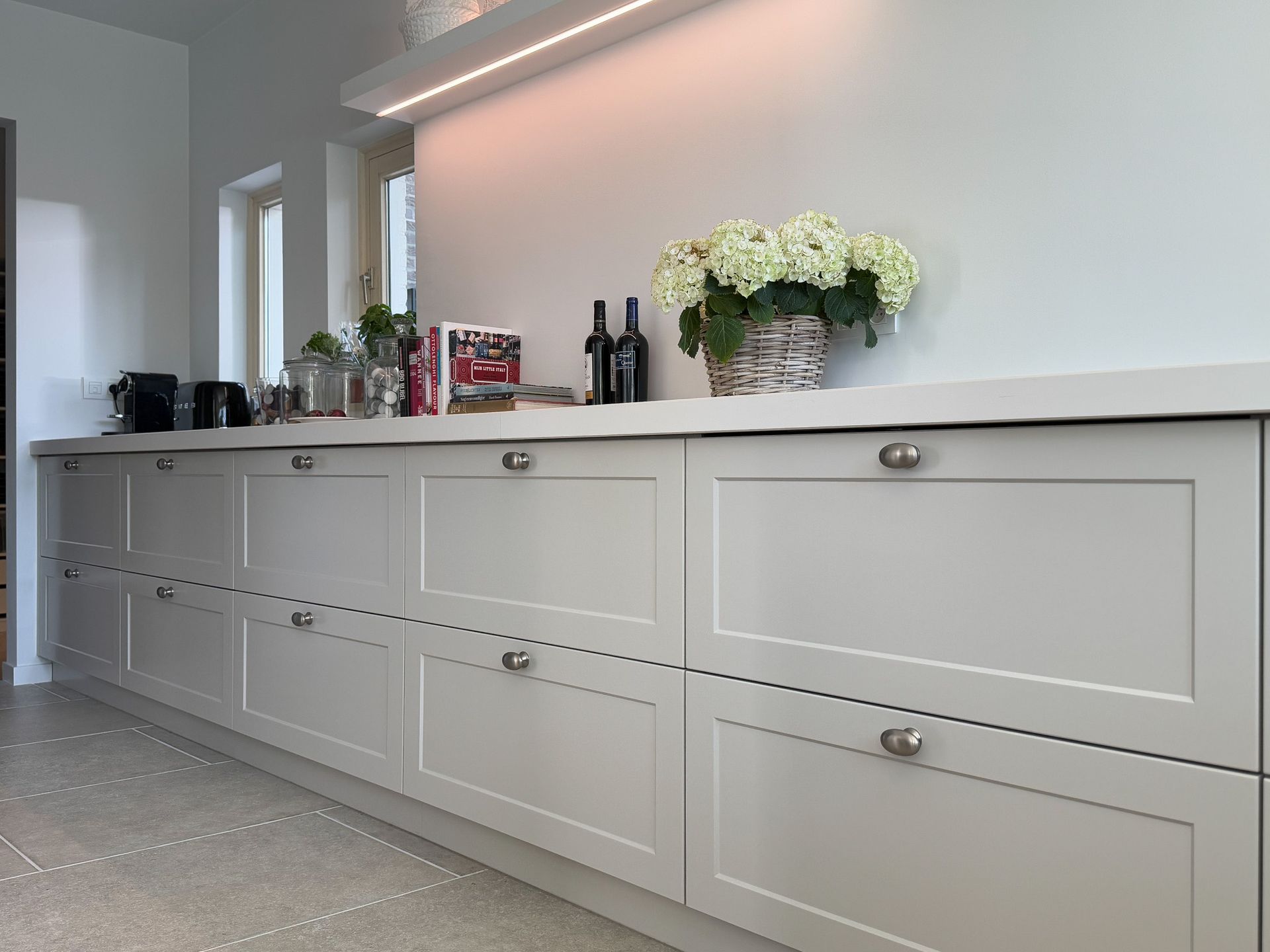 Lichtgrijze keukenkastjes met een wit aanrechtblad, bloemen, flessen en apparaten tegen een witte muur.