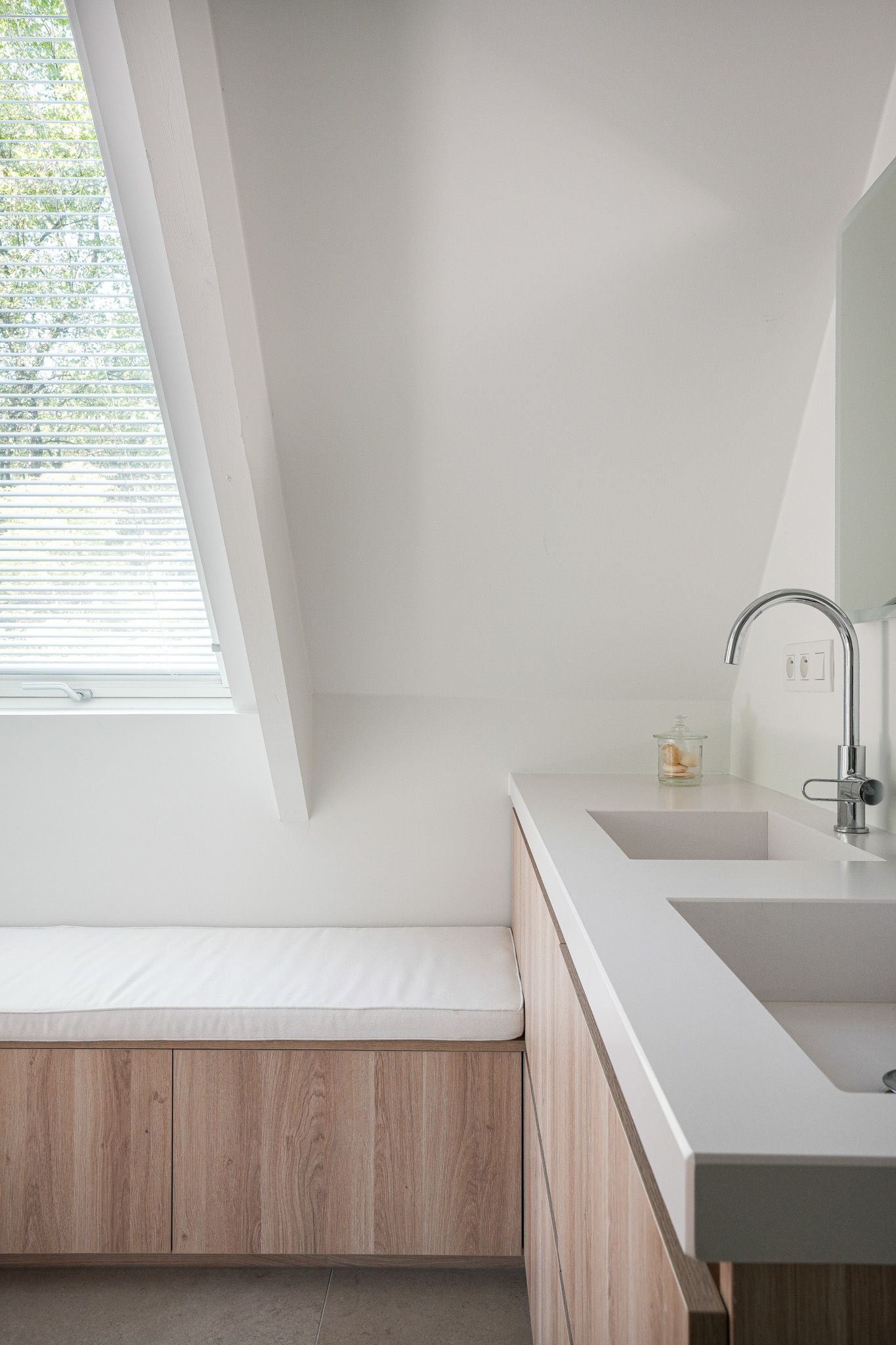 Badkamer met houten kasten, witte aanrechtbladen, schuin plafond en raam met jaloezieën.