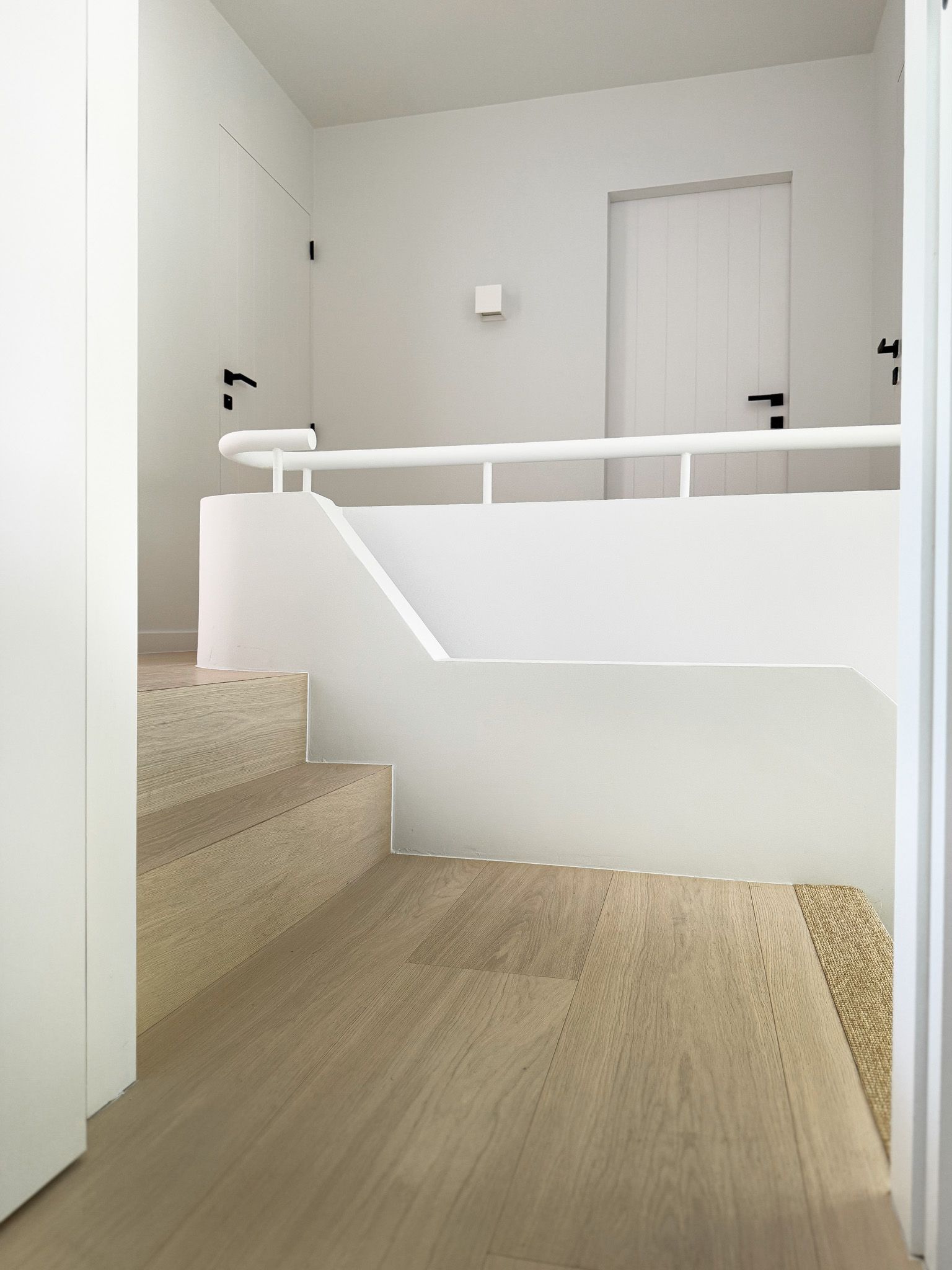 Witte trap met houten treden en leuning. Deuren met zwarte handgrepen op de achtergrond.