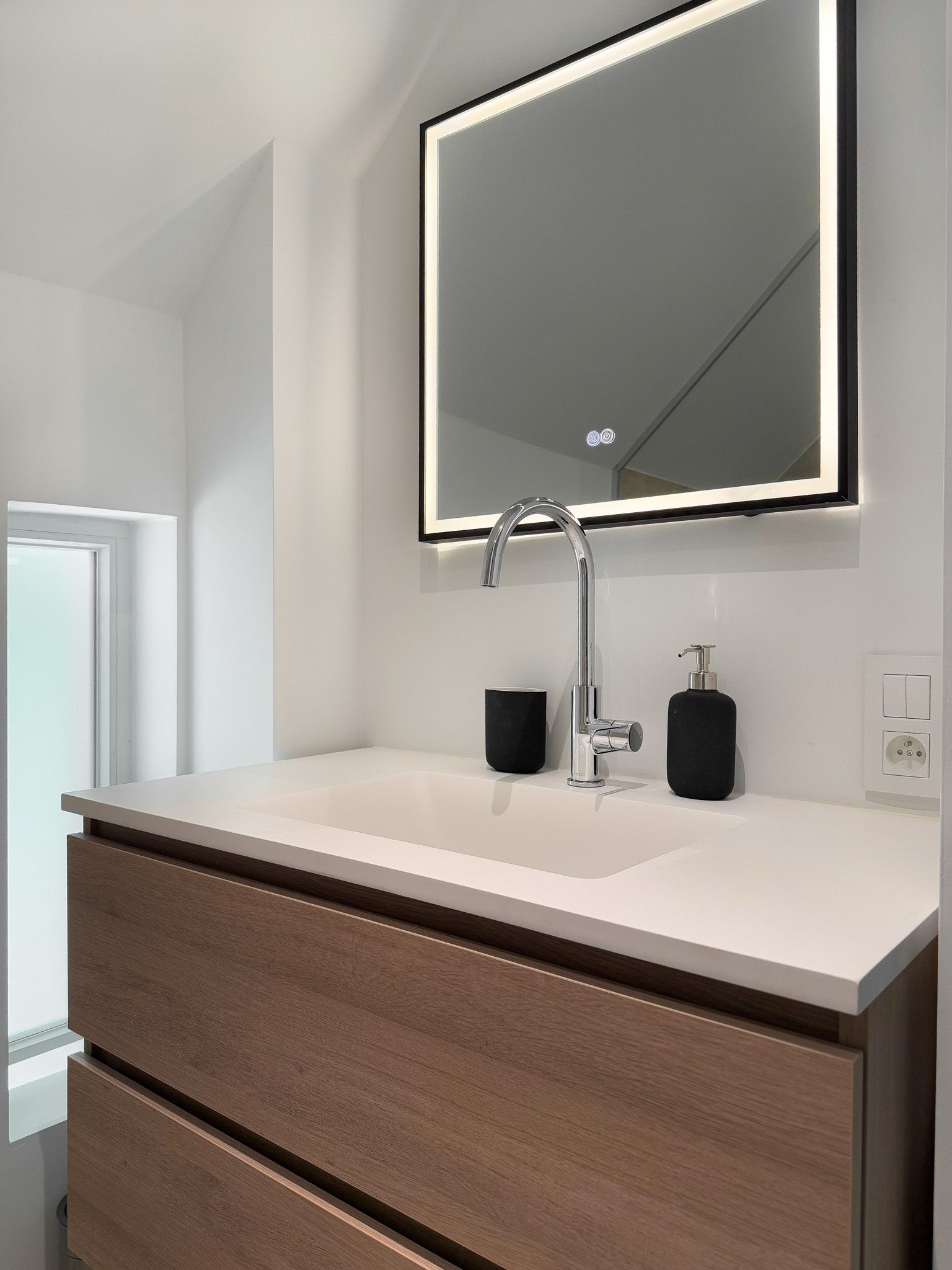 Moderne badkamer met houten wastafelmeubel, wit aanrechtblad, verlichte spiegel en chromen kraan.