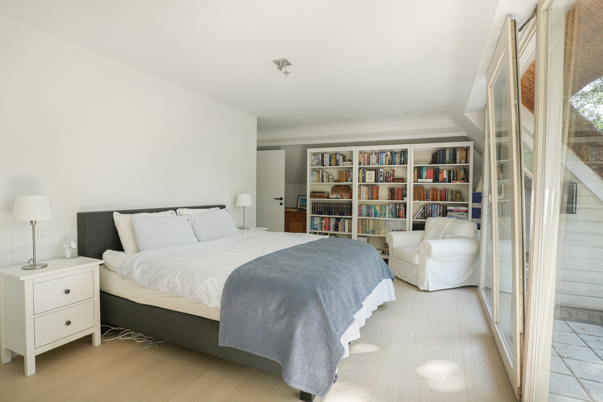 Slaapkamer met bed, witte muren, boekenkast en open glazen deuren naar een terras.