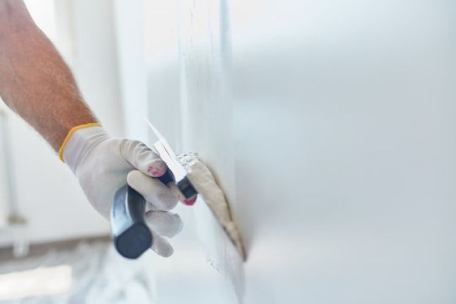 Persoon met witte handschoenen gebruikt een troffel om stopverf op een witte muur aan te brengen.