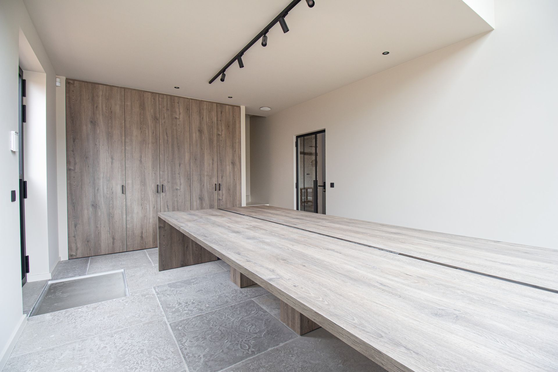 Lege, moderne kamer met een lange houten tafel, ingebouwde houten kasten en een zwarte glazen deur.