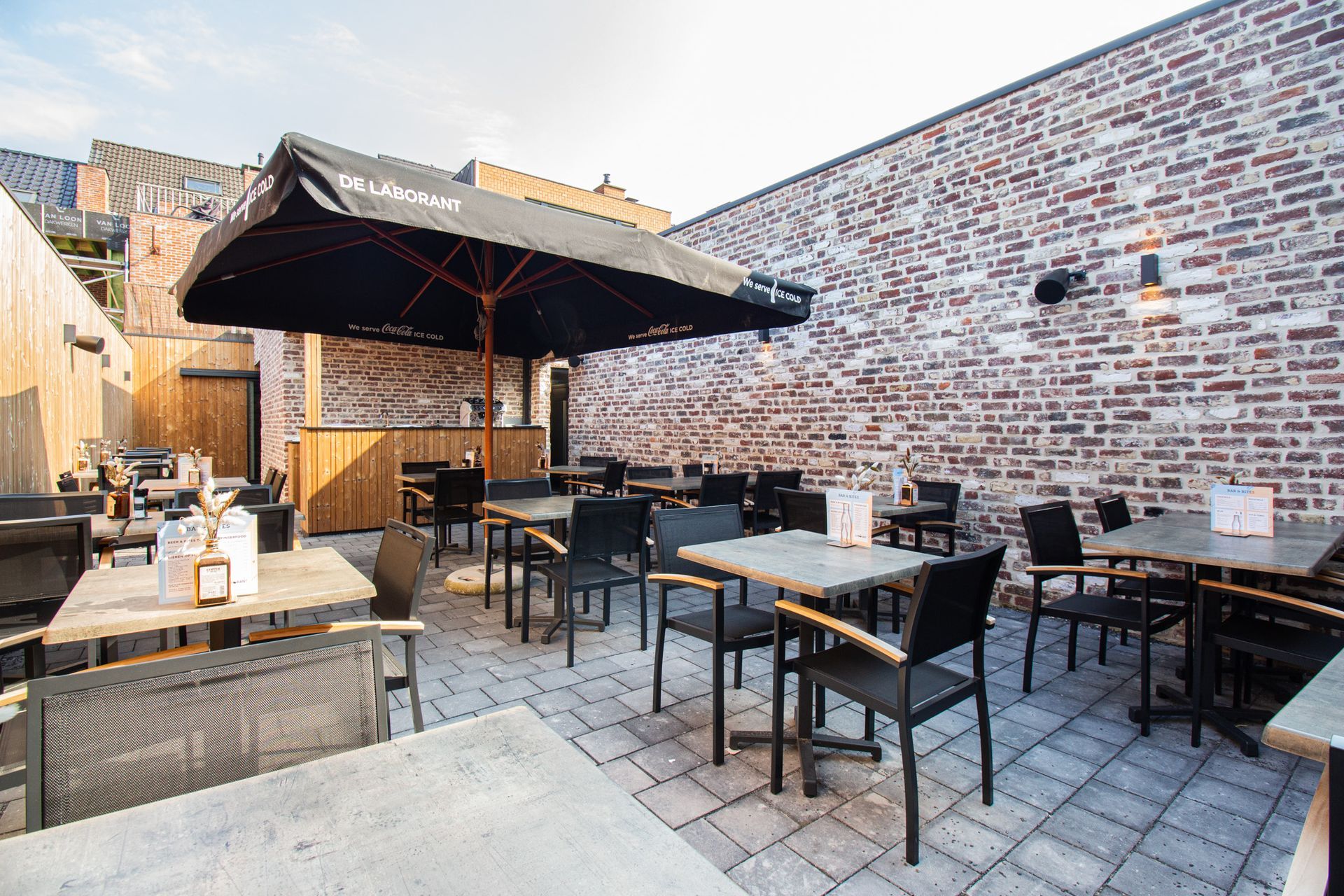 Terras met tafels, stoelen en een grote parasol tegen een bakstenen muur.