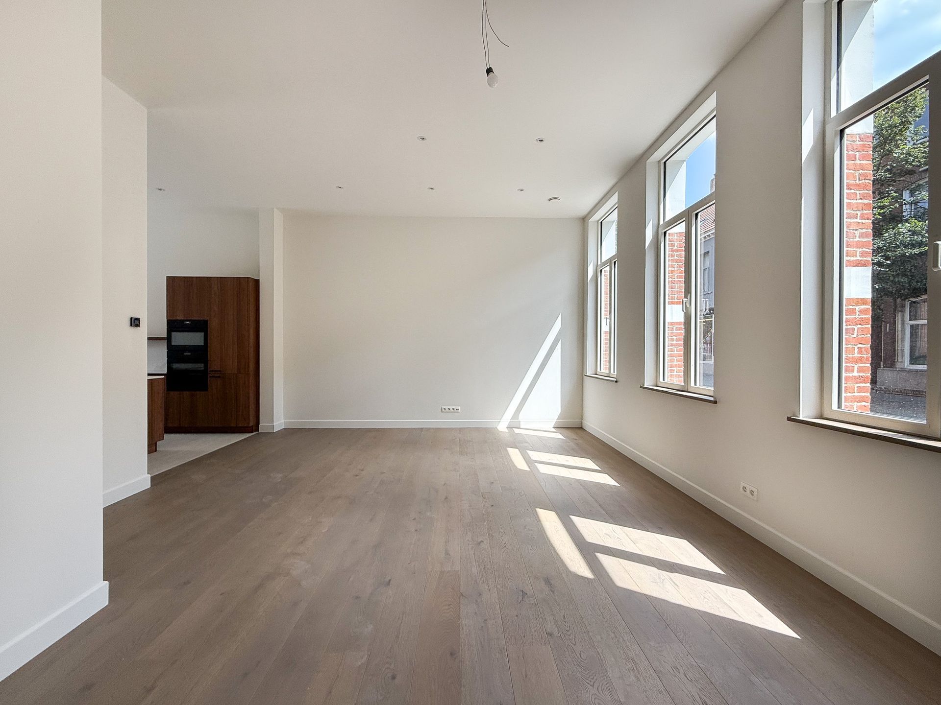 Lege, lichte kamer met houten vloeren, hoge ramen en een kleine keuken.