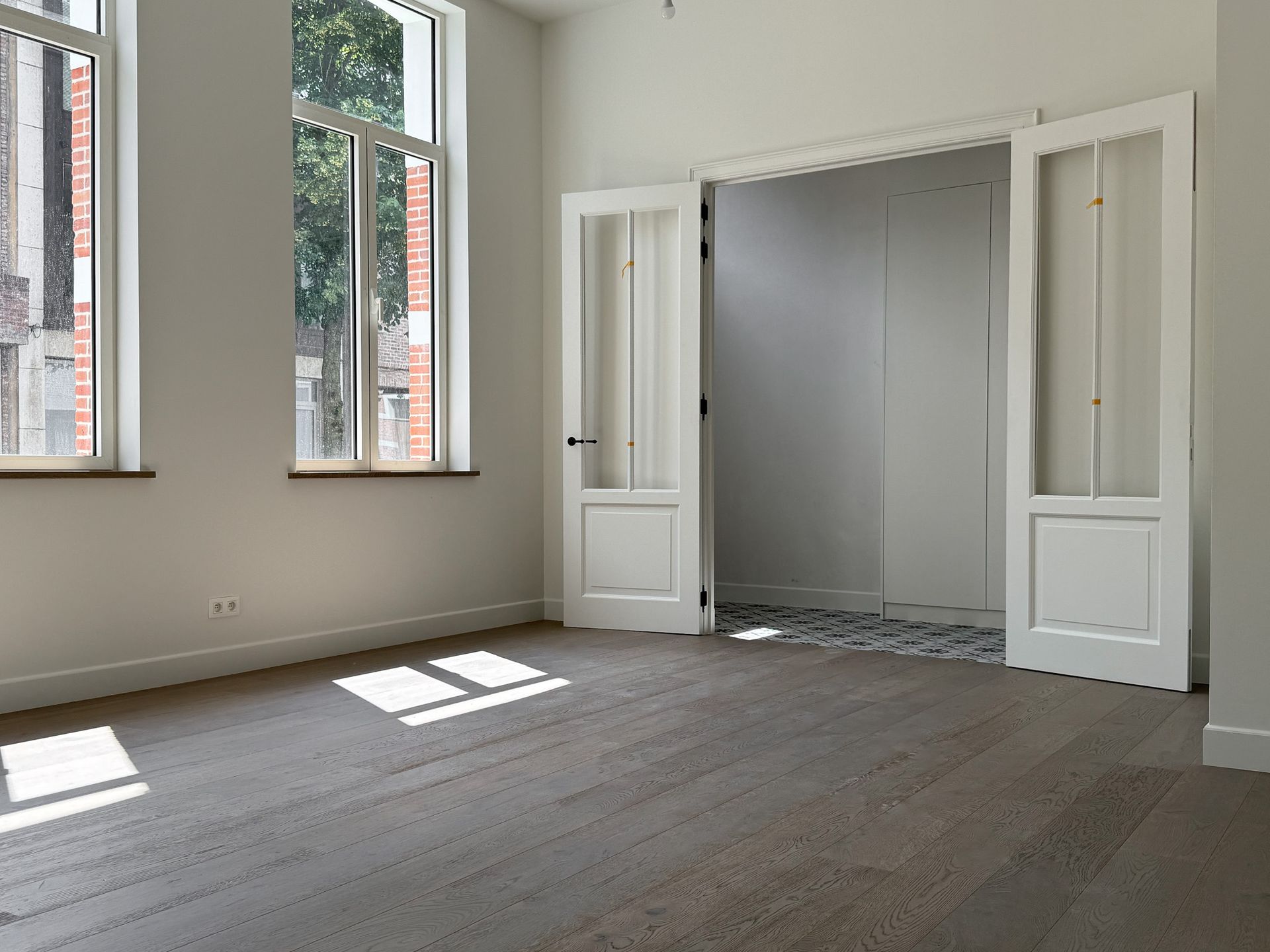 Lege kamer met houten vloer, hoge ramen en witte dubbele deuren die naar een andere ruimte leiden.