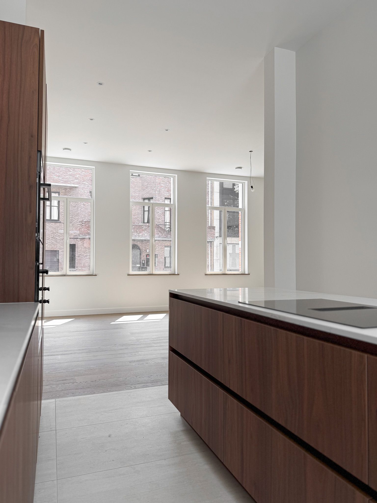 Moderne keuken met houten kasten, witte aanrechtbladen en grote ramen met uitzicht op een bakstenen gebouw.