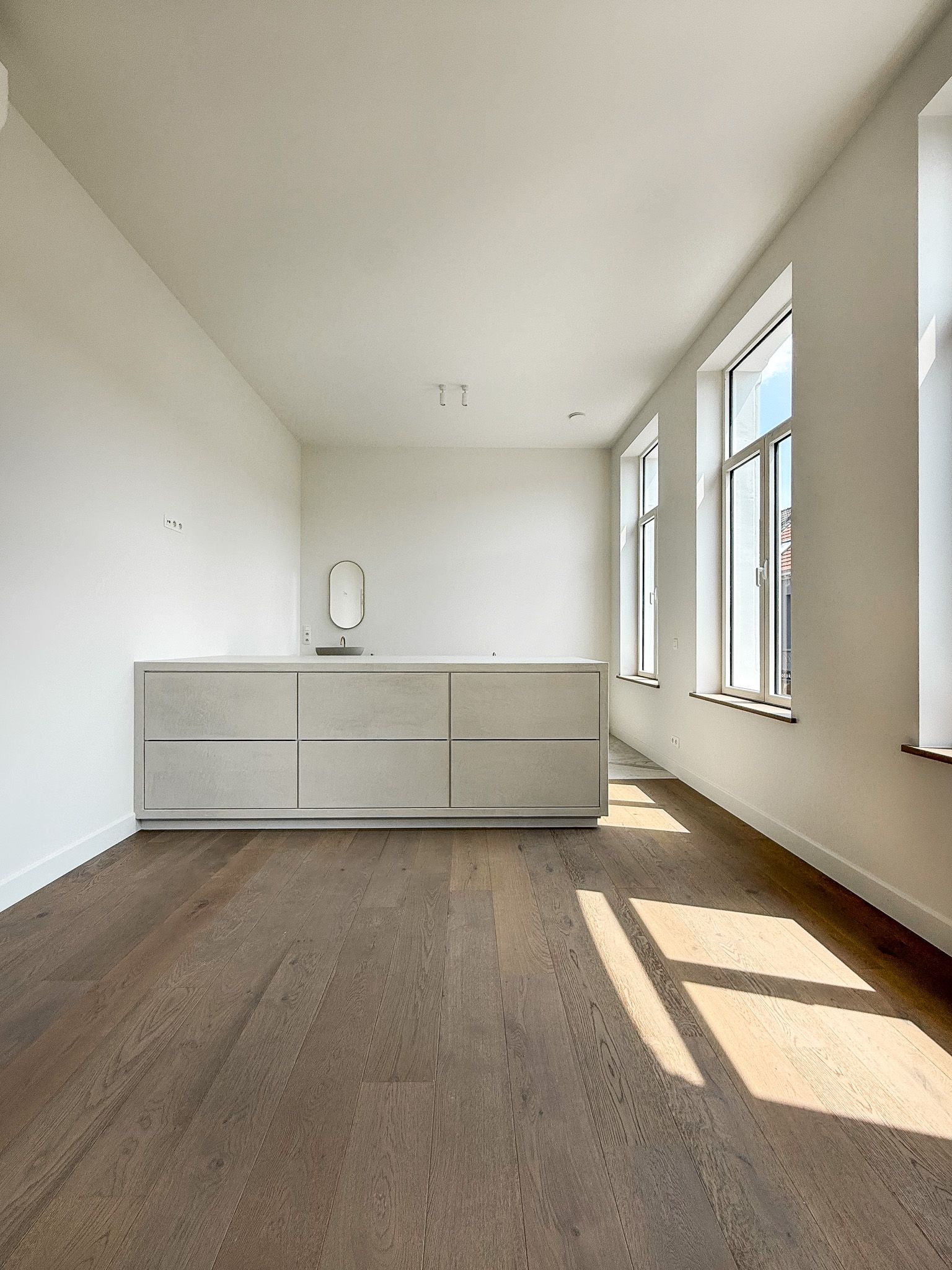 Lege, lichte kamer met houten vloeren, witte muren, ingebouwde ladekast en ramen die schaduwen werpen.
