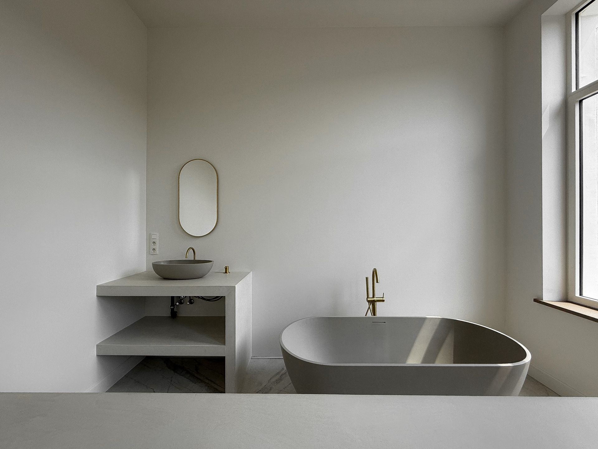 Minimalistische badkamer: grijs bad, wastafel en wastafel. Gouden kraan en ovale spiegel. Raam rechts.
