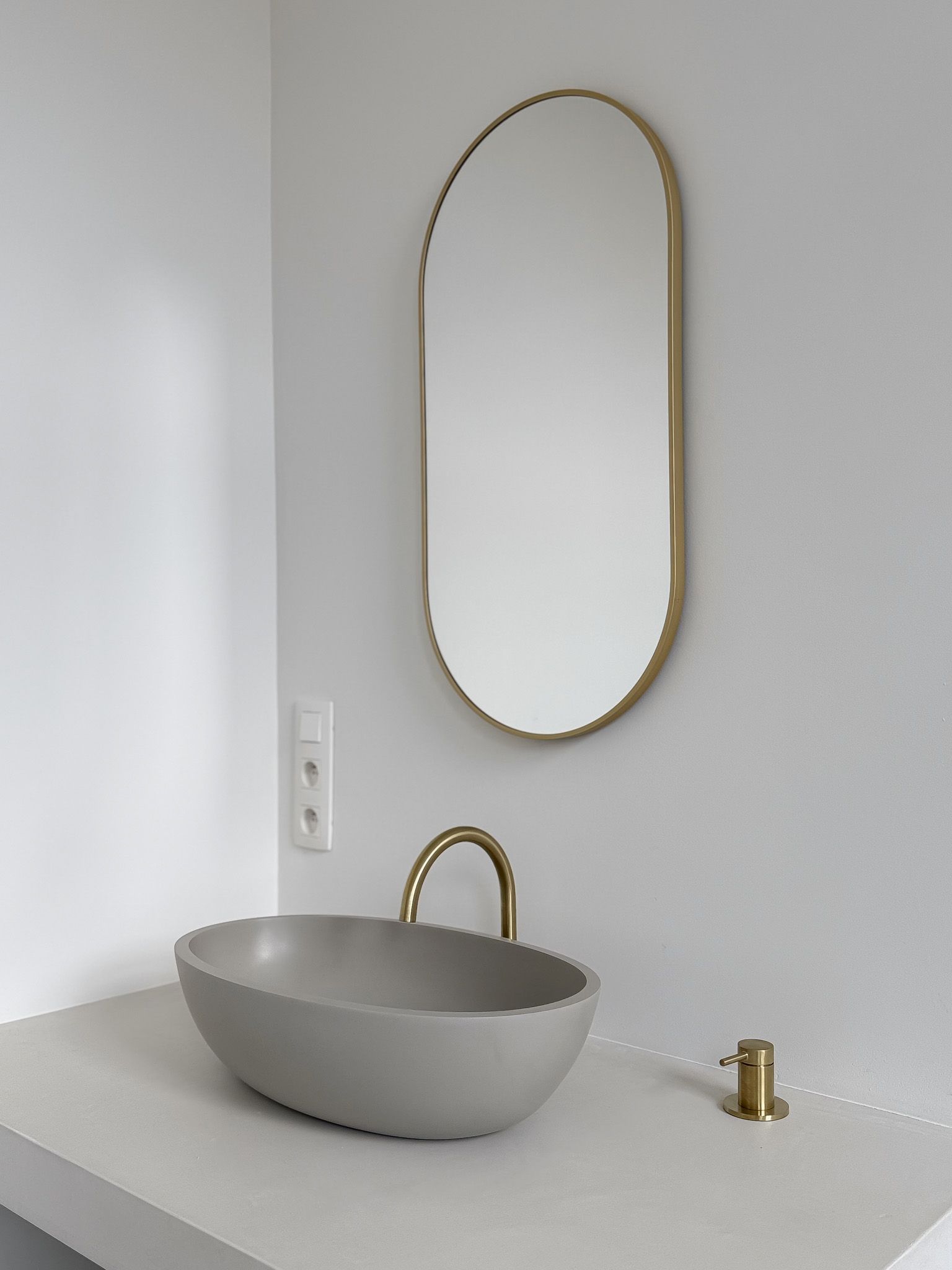 Ovale spiegel met gouden lijst boven grijze ovale wastafel en gouden kraan in een witte badkamer.